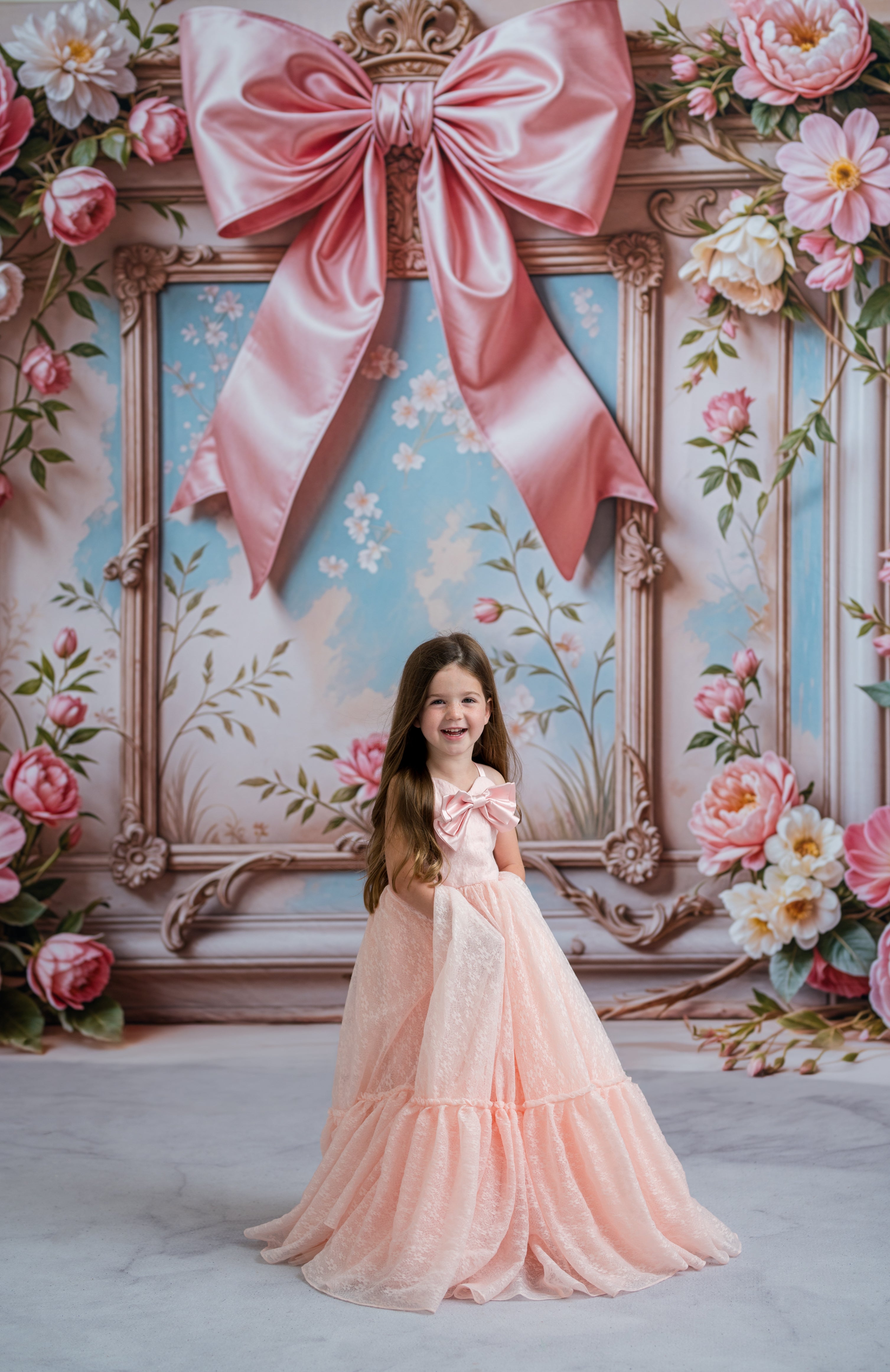 Young girl in a pink dress standing in front of a decorative wall with a large pink bow and floral decorations.