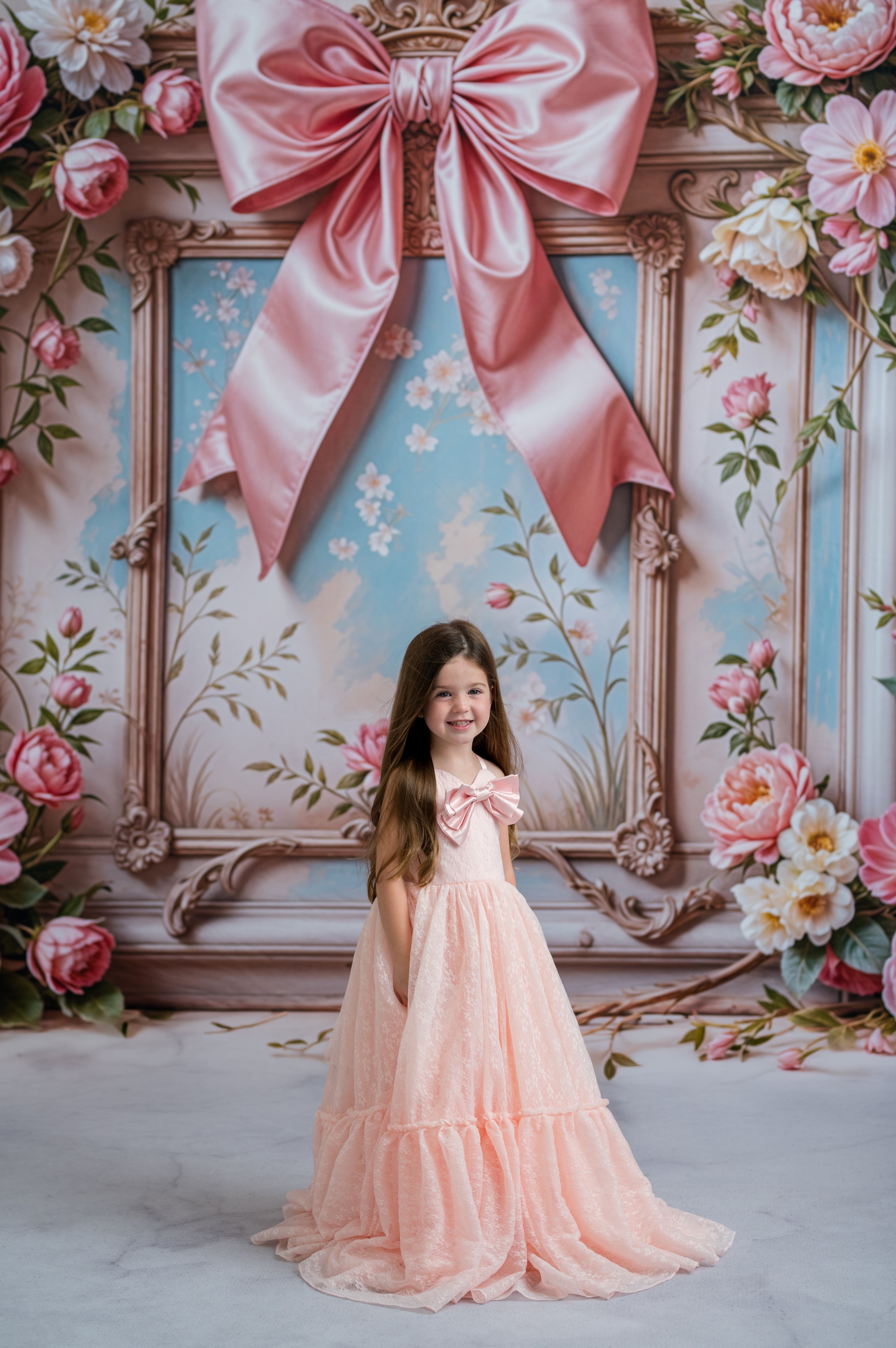 Young girl in a pink dress standing in front of a decorative wall with a large pink bow and floral arrangements.