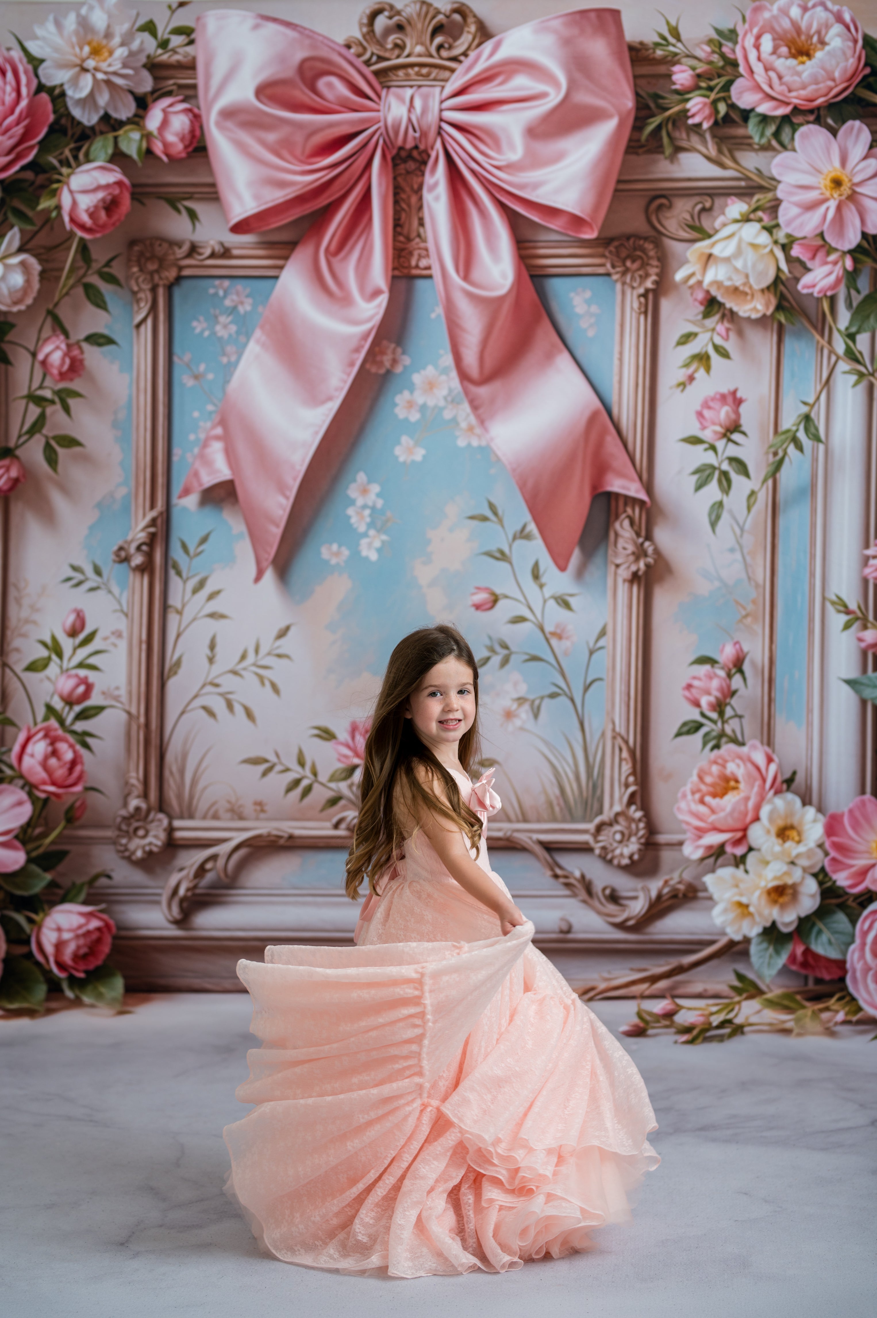 Young girl in a pink dress standing in front of a decorative backdrop with flowers and a large bow.