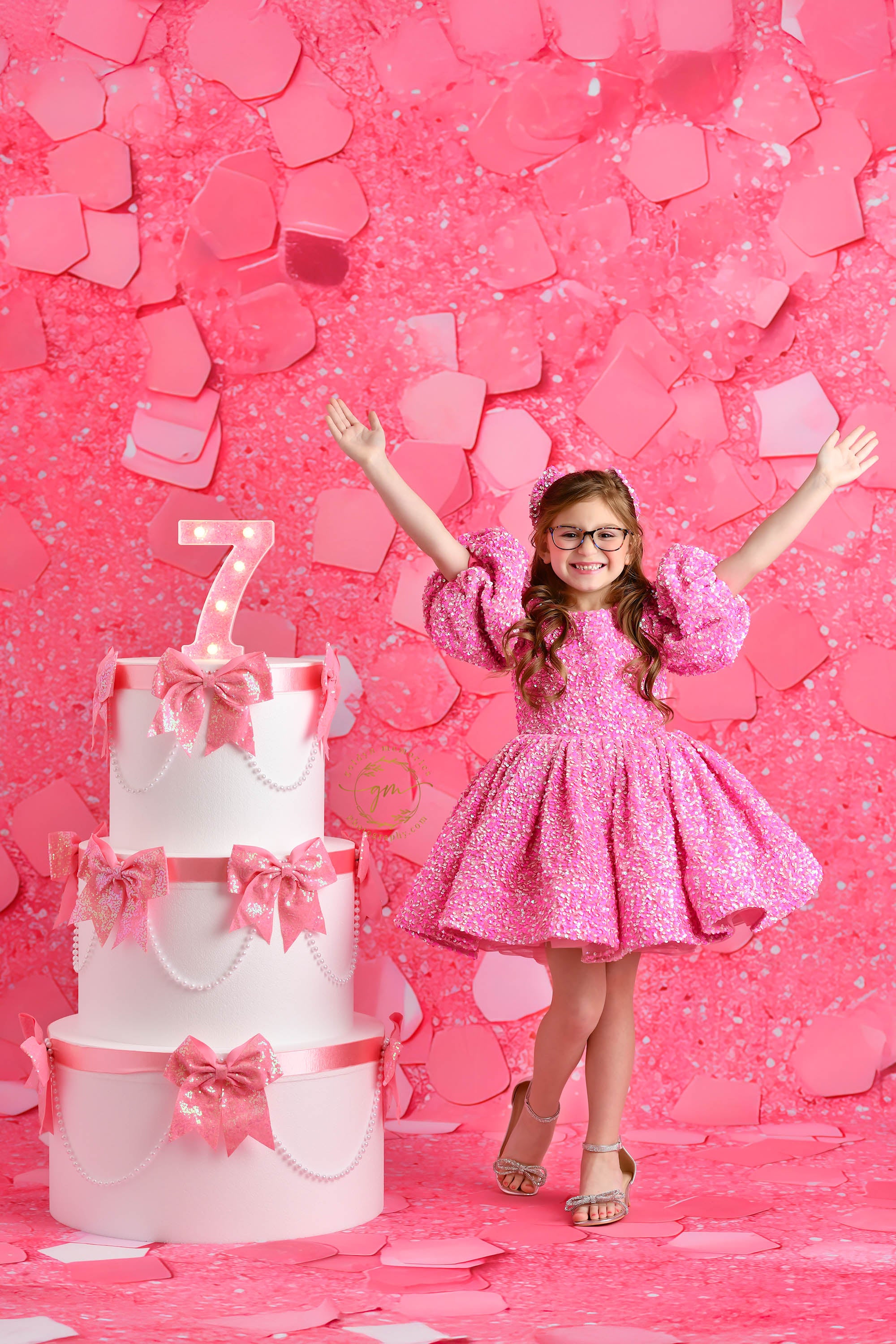 A beaming child in a glittery pink dress twirls with excitement in front of a pink confetti wall, standing next to an elegant, tiered cake adorned with pink bows and a light-up "7" sign.