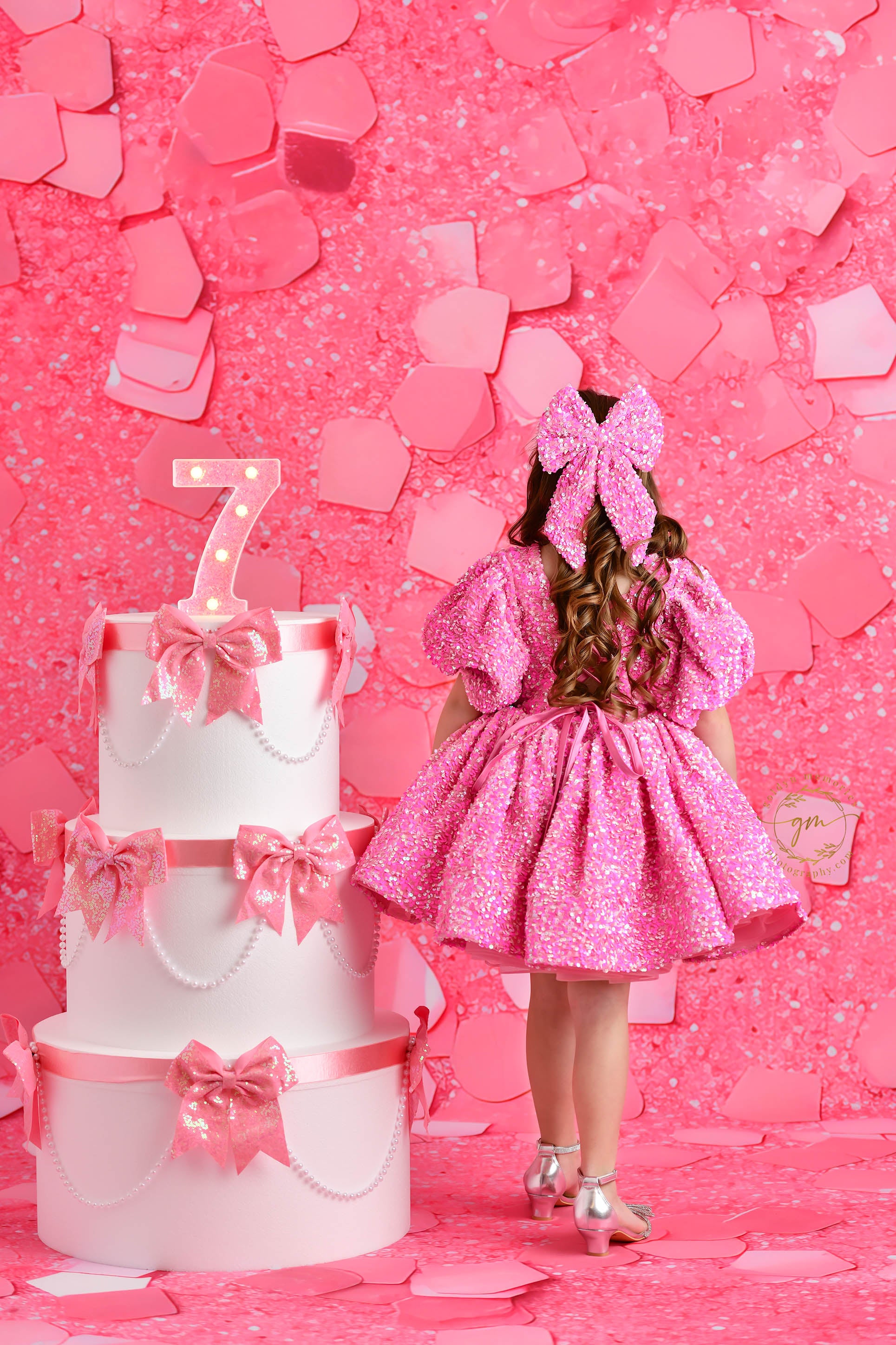 A joyful young girl in a sparkly pink dress with puff sleeves stands with her arms raised in front of a pink confetti-themed backdrop, smiling brightly next to a decorated birthday cake with a glowing number "7" topper.