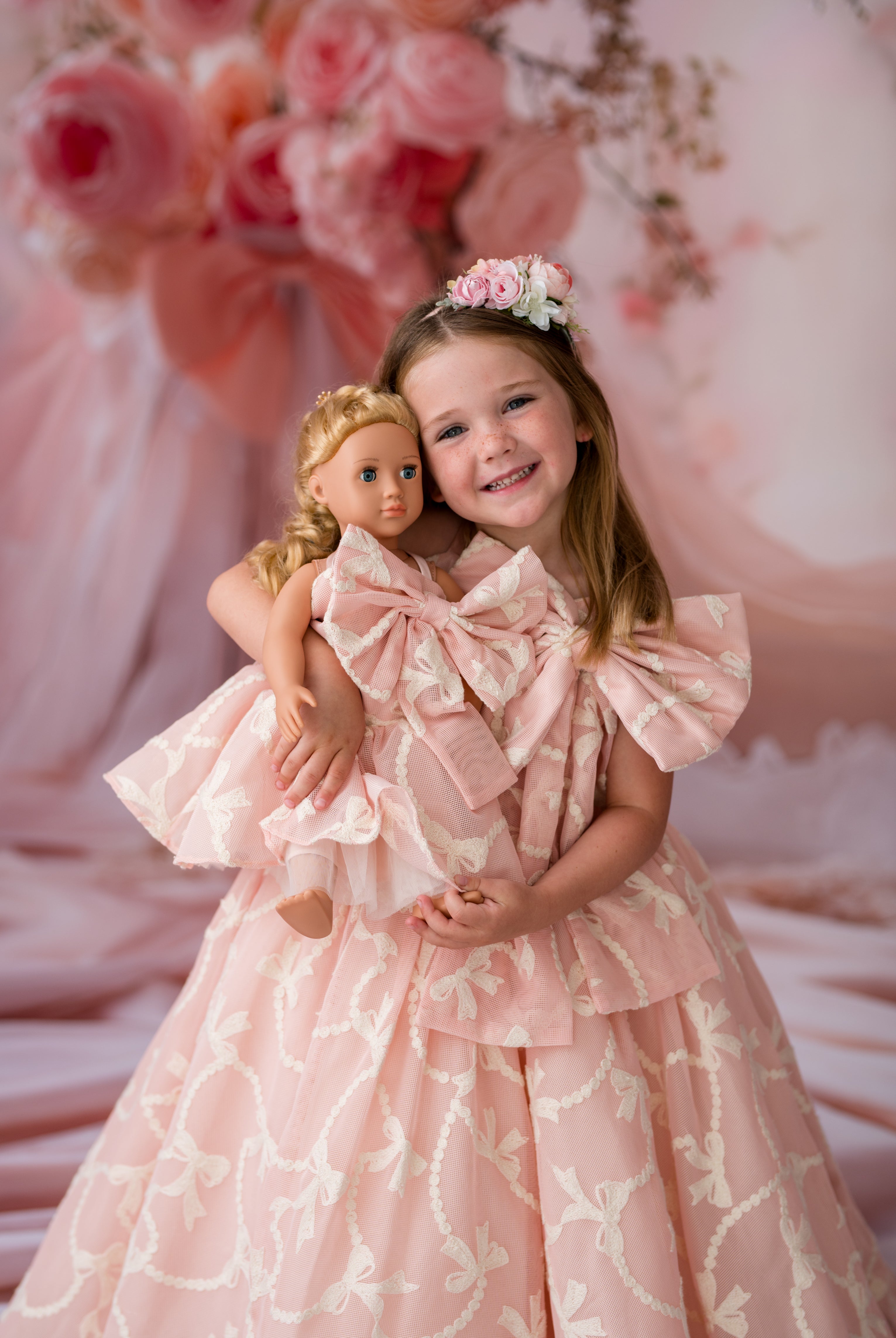 Young girl in a pink dress holding a doll against a floral backdrop
