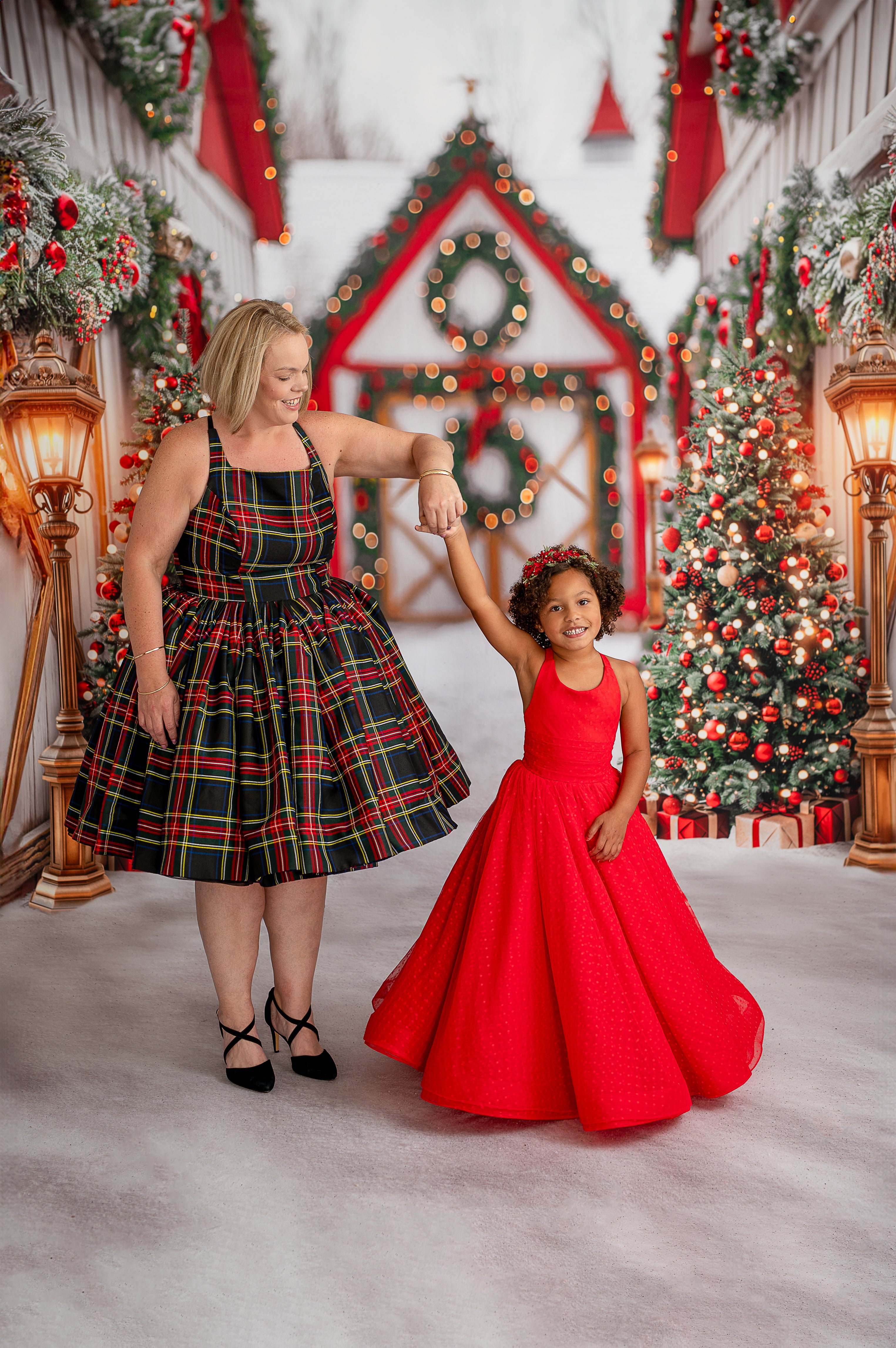 "The image shows a woman in a plaid dress joyfully holding her daughter in a red frock against a rich red background, decorated with Christmas trees and a shelf filled with colorful presents and holiday decorations."