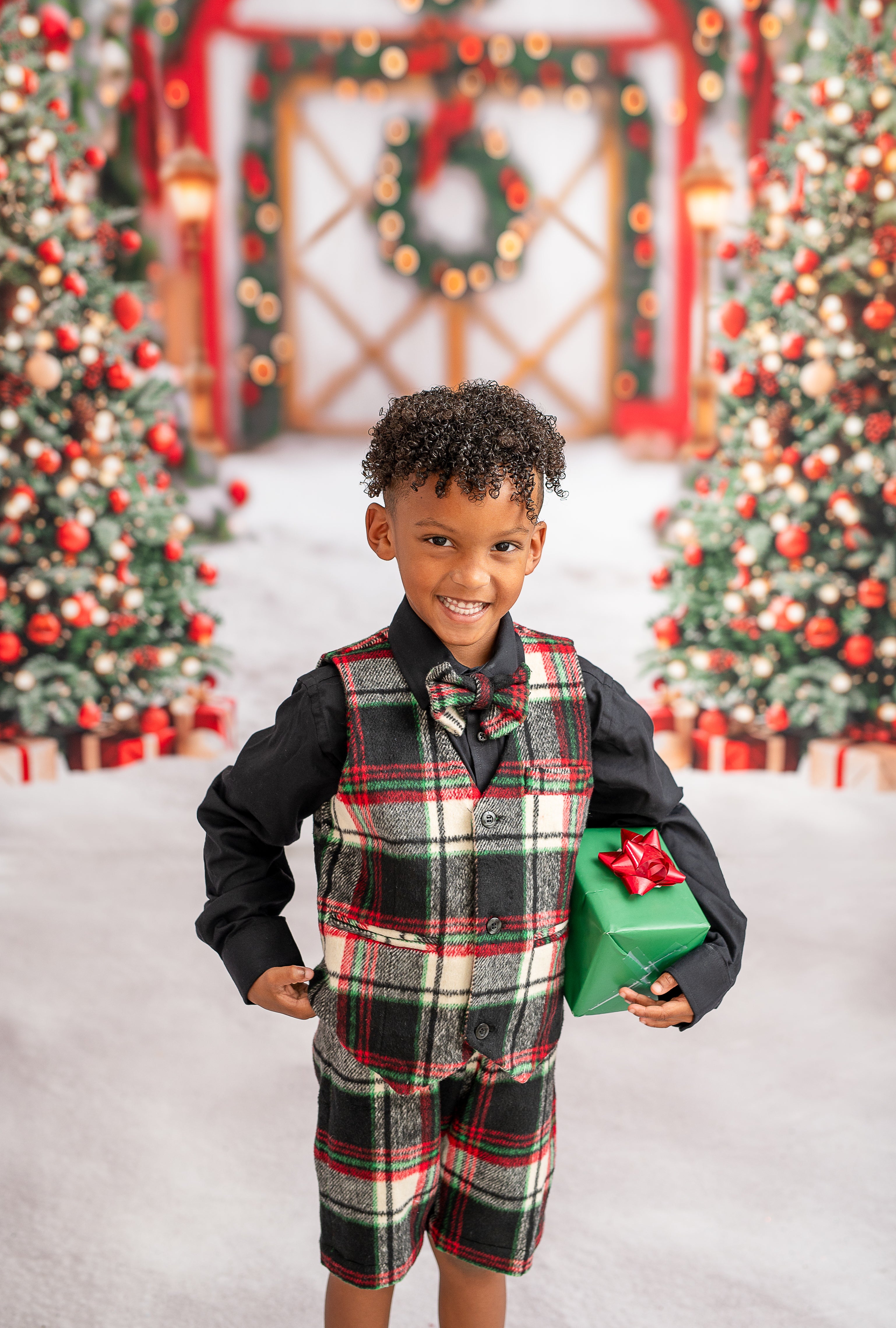 "A smiling boy wearing a plaid outfit, including a vest and shorts, poses with a wrapped gift in front of a holiday backdrop. The scene features lit Christmas trees and a warmly decorated archway, enhancing the festive atmosphere."