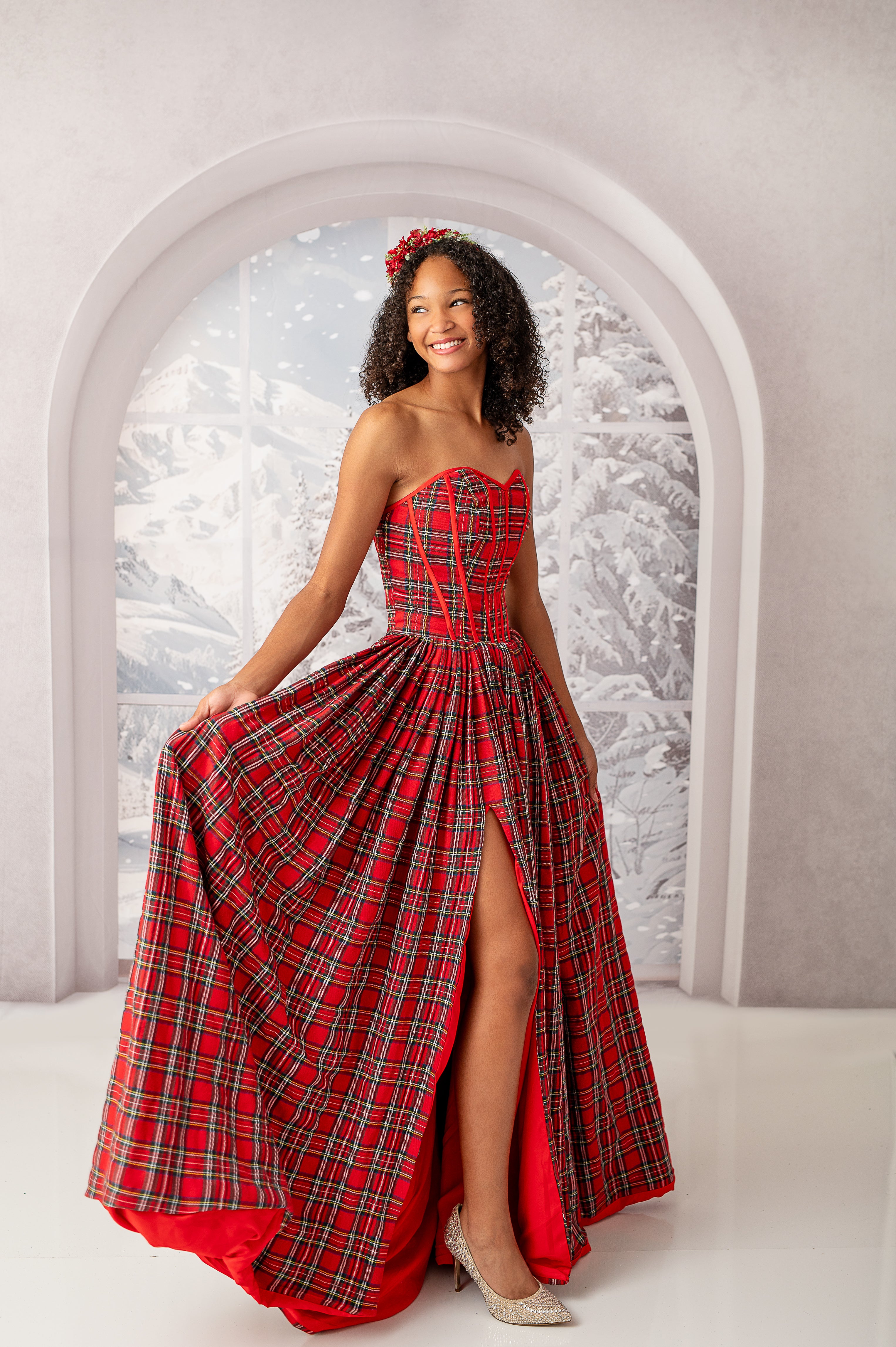 "A cheerful young woman showcases a red and green plaid gown with a sweetheart neckline and floor-length skirt with a front slit. The backdrop is an arched window with a view of a snow-filled, winter wonderland."