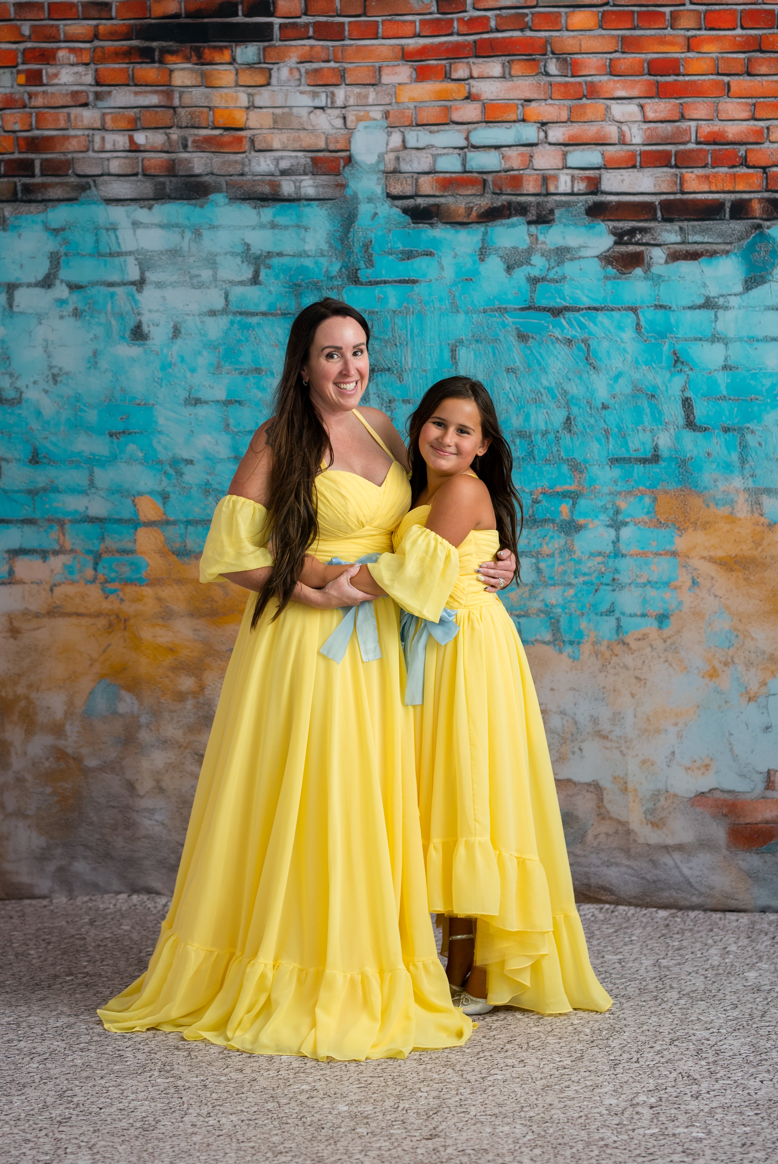 Two women in yellow dresses standing against a colorful brick wall.