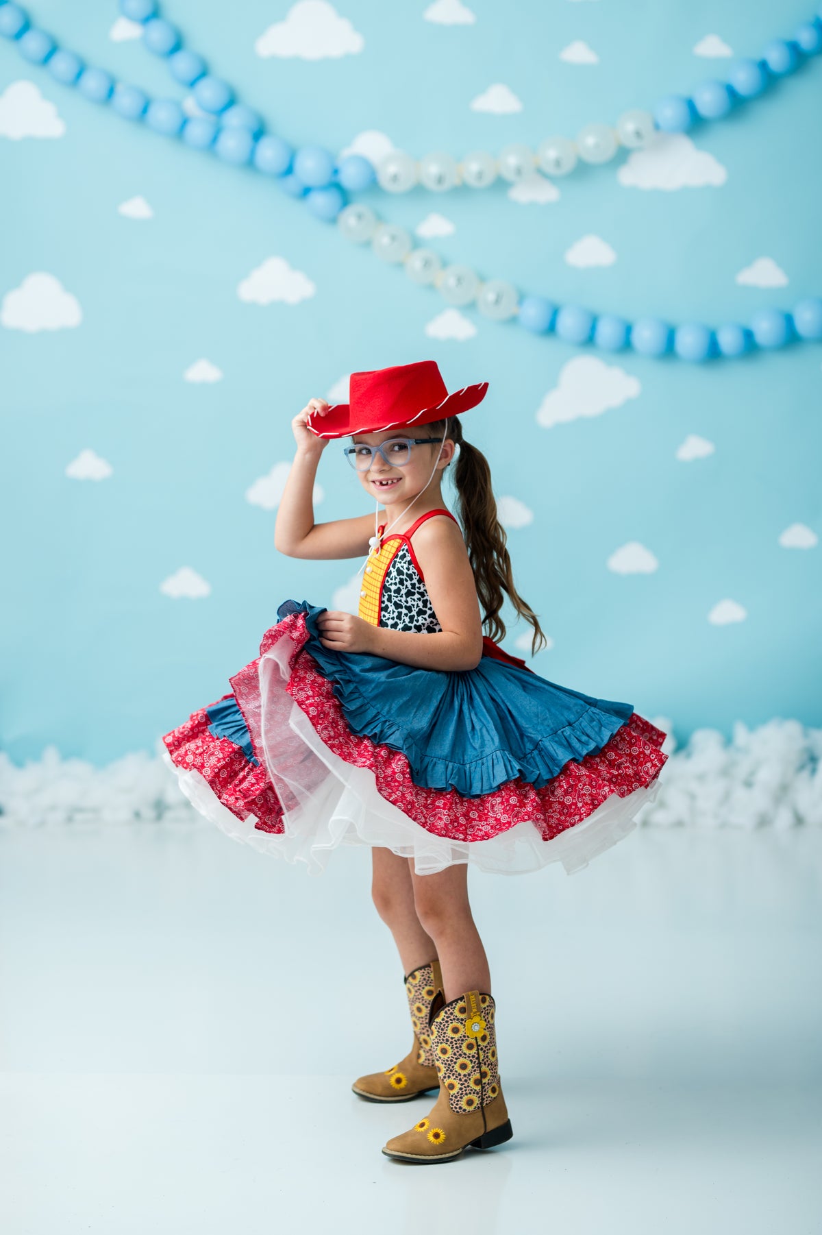 A young girl modeling a colorful cowgirl costume consisting of a red cowboy hat, a blue ruffled skirt with red and white trim, a patterned top, and yellow western boots, set against a light blue backdrop decorated with clouds and bead garlands.