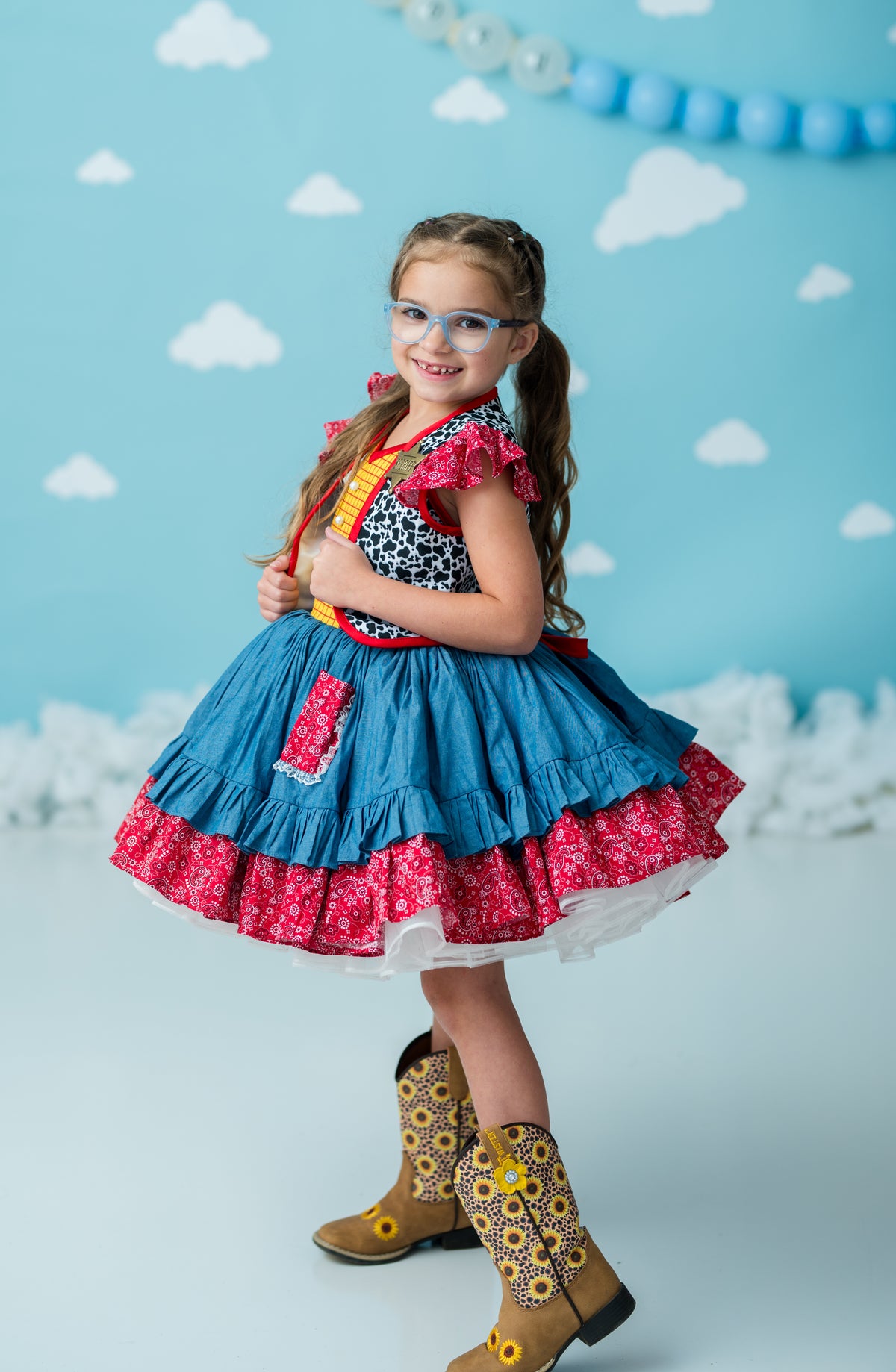 A young girl with glasses and a ponytail stands in a studio wearing a blue and pink tiered dress with a black-and-white patterned bodice, red floral ruffle trim, and tan cowboy boots against a light blue cloud-patterned backdrop.
