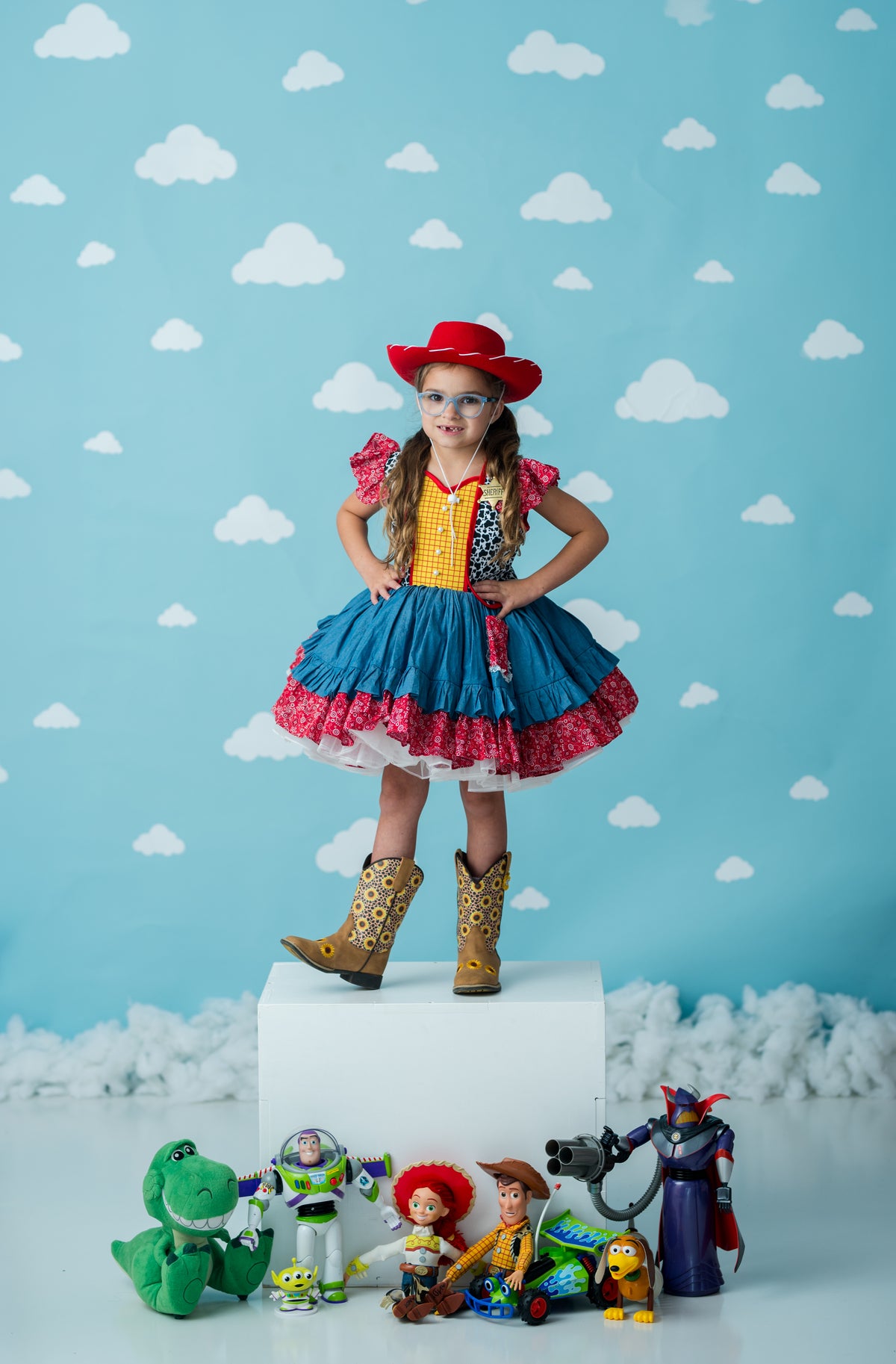 A young girl stands on a white pedestal wearing a Woody-inspired costume with a red cowboy hat, yellow vest, blue skirt, and brown boots. Toy characters and plush figures are arranged at her feet against a light blue backdrop with white cloud patterns.