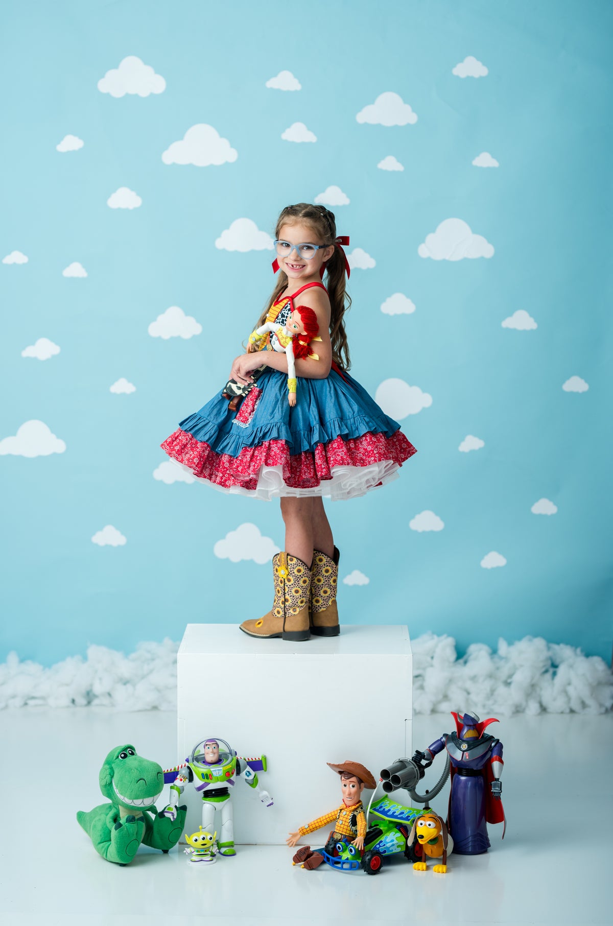 A young girl stands on a white pedestal wearing a blue, red, and white ruffled costume dress with cowboy boots, holding a plush toy. In front of her are various Toy Story character figurines against a light blue backdrop with white cloud patterns.
