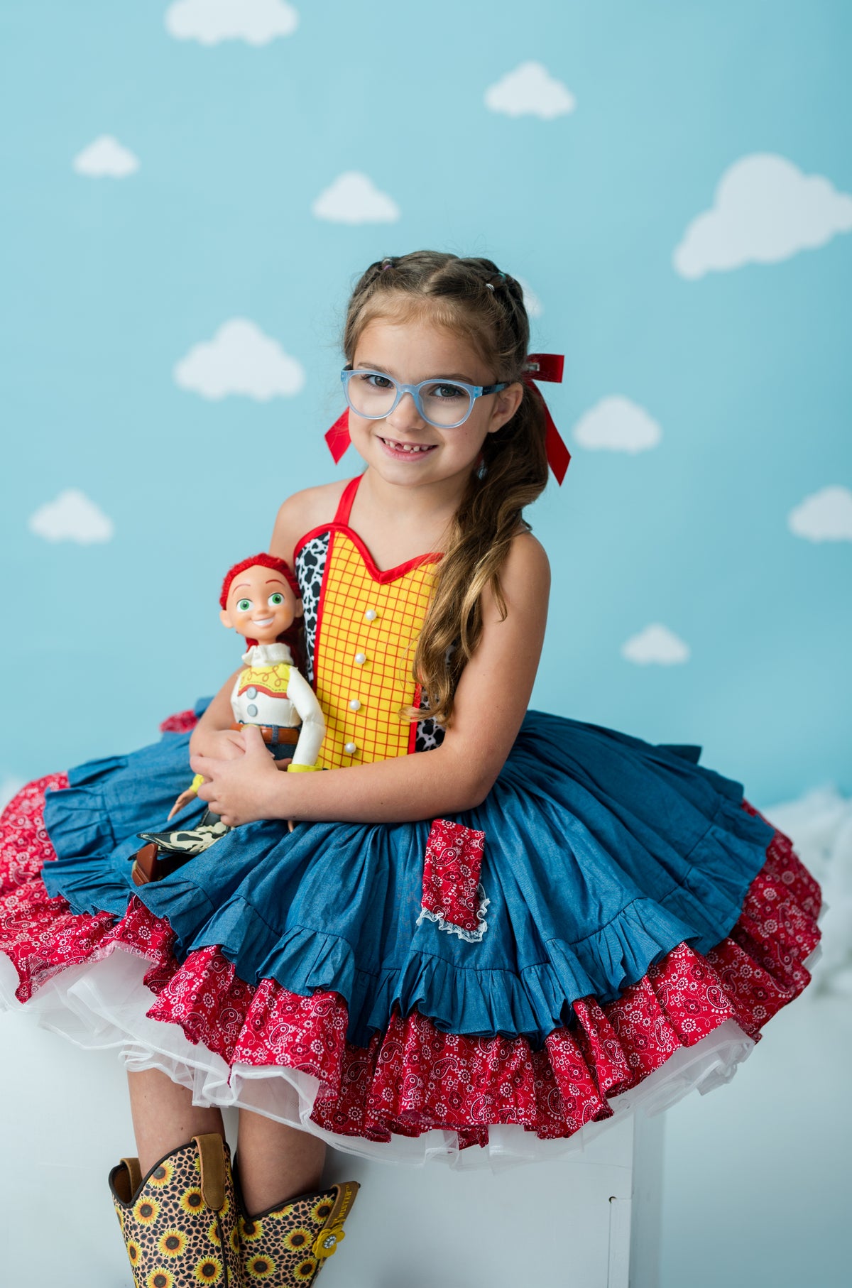 A young girl with glasses and red hair bows sits on a white stool wearing a colorful costume dress inspired by Toy Story, featuring a yellow gingham top, a blue ruffled skirt with red patterned trim, and yellow cowboy boots. She holds a doll against a light blue background with white cloud illustrations.