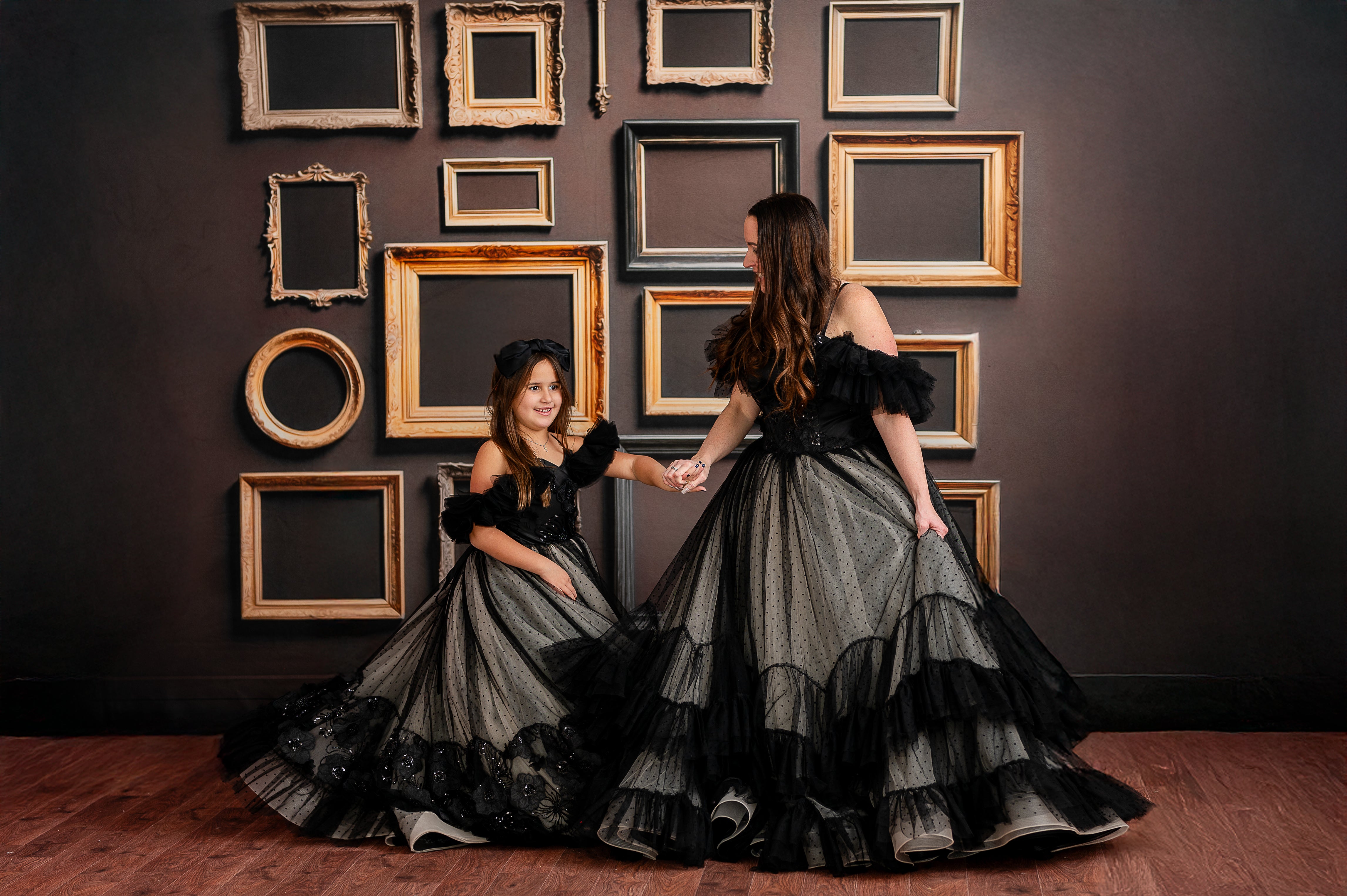 A model in a black lace-adorned gown stands poised in front of an artistic frame wall.