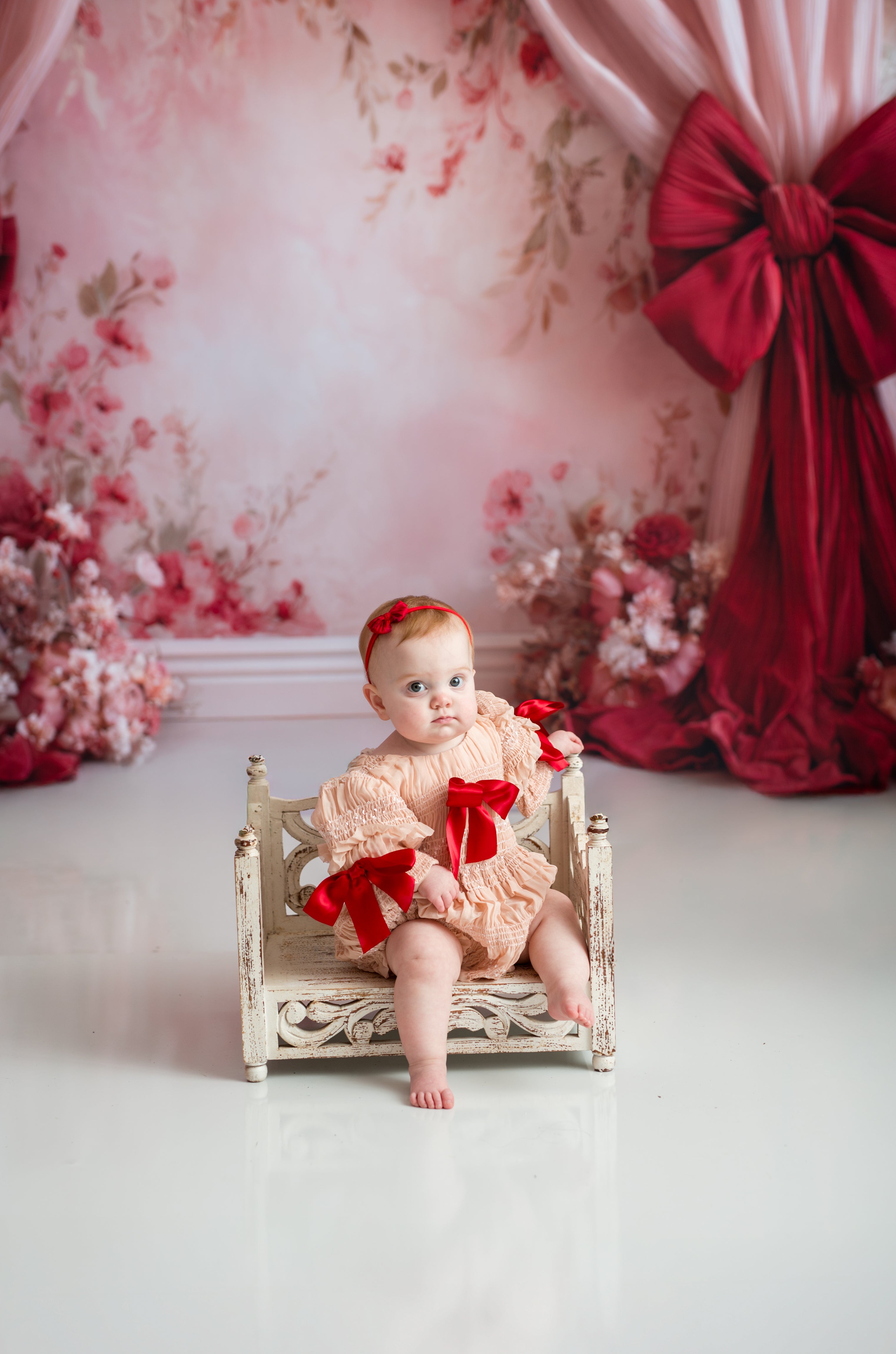 Baby in a pink outfit with red bows sitting on a small bed against a floral backdrop