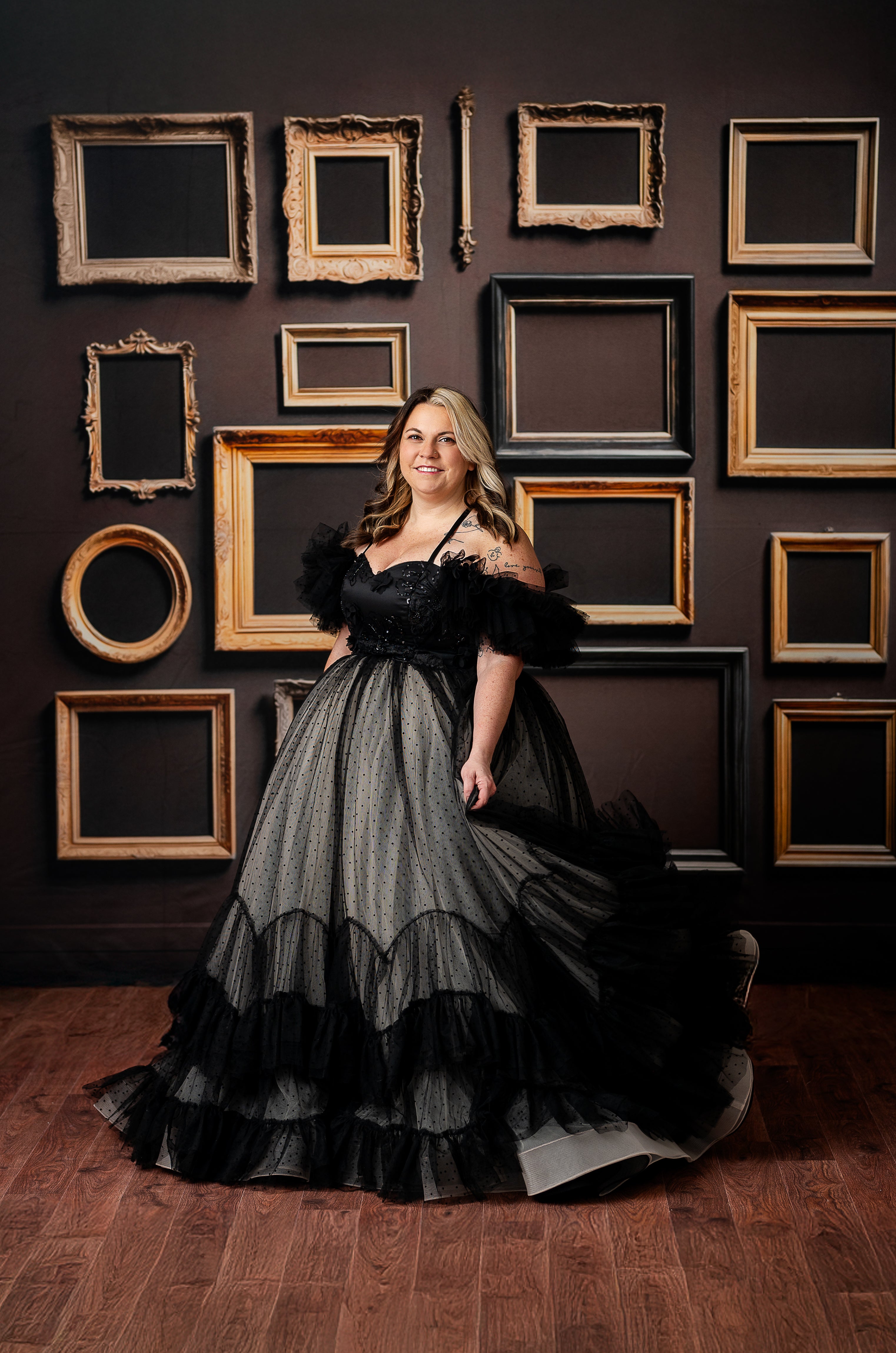 A woman in a black and nude polka-dot tulle gown poses gracefully against a wall of empty vintage frames.