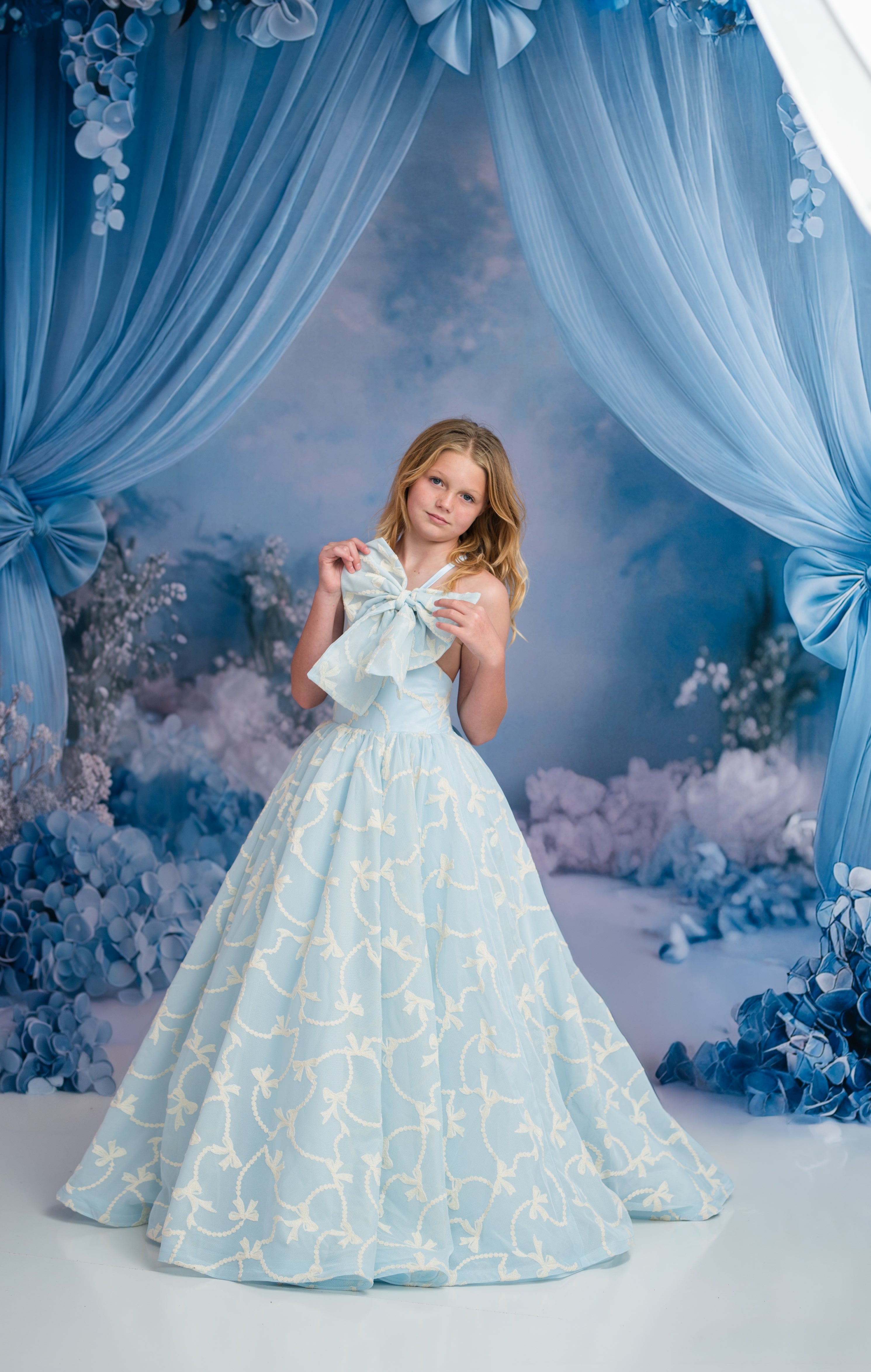Woman in a light blue gown with floral patterns standing in front of decorative blue curtains and floral arrangements.