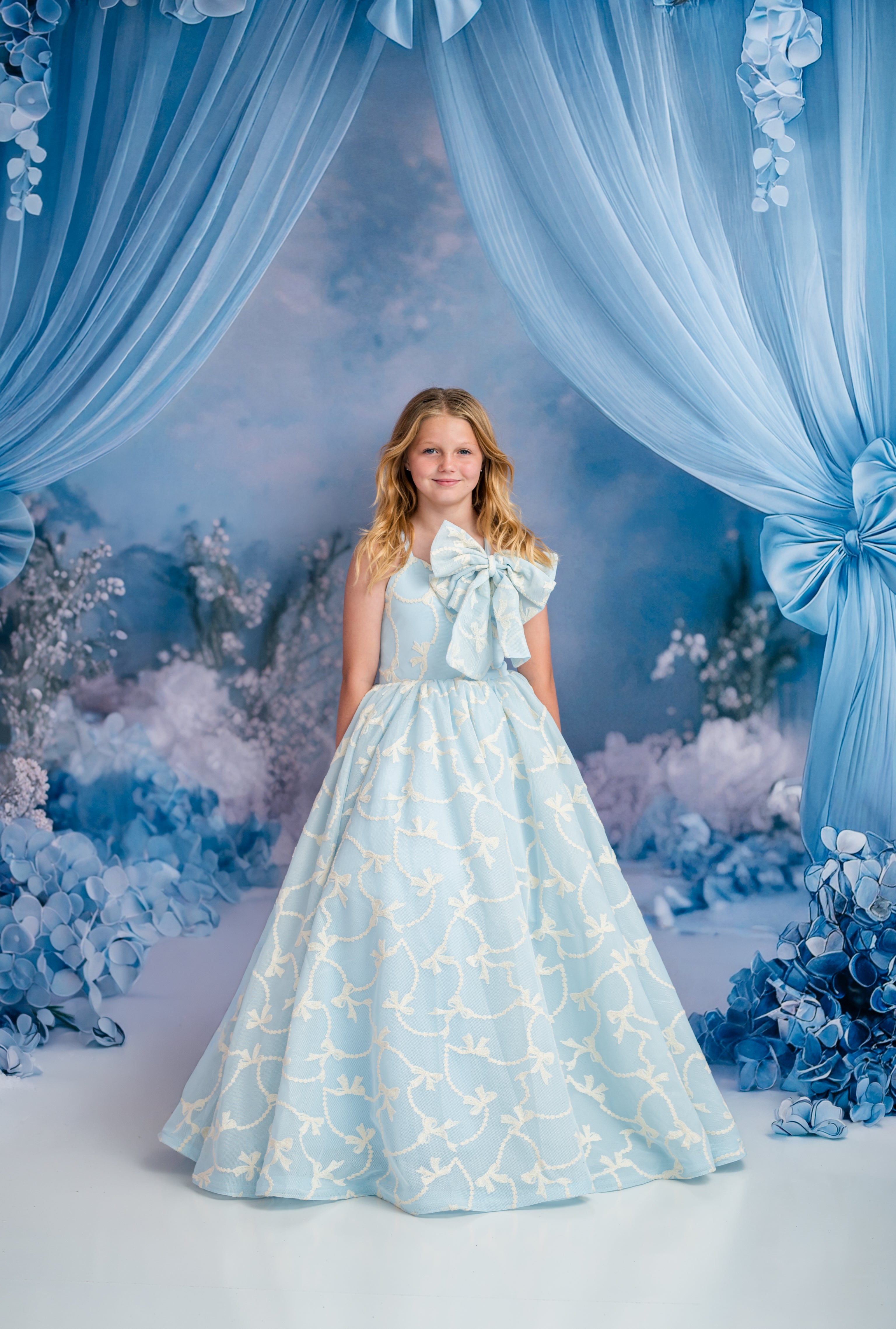 Young girl in a light blue dress with bow details standing in front of blue floral and curtain backdrop.