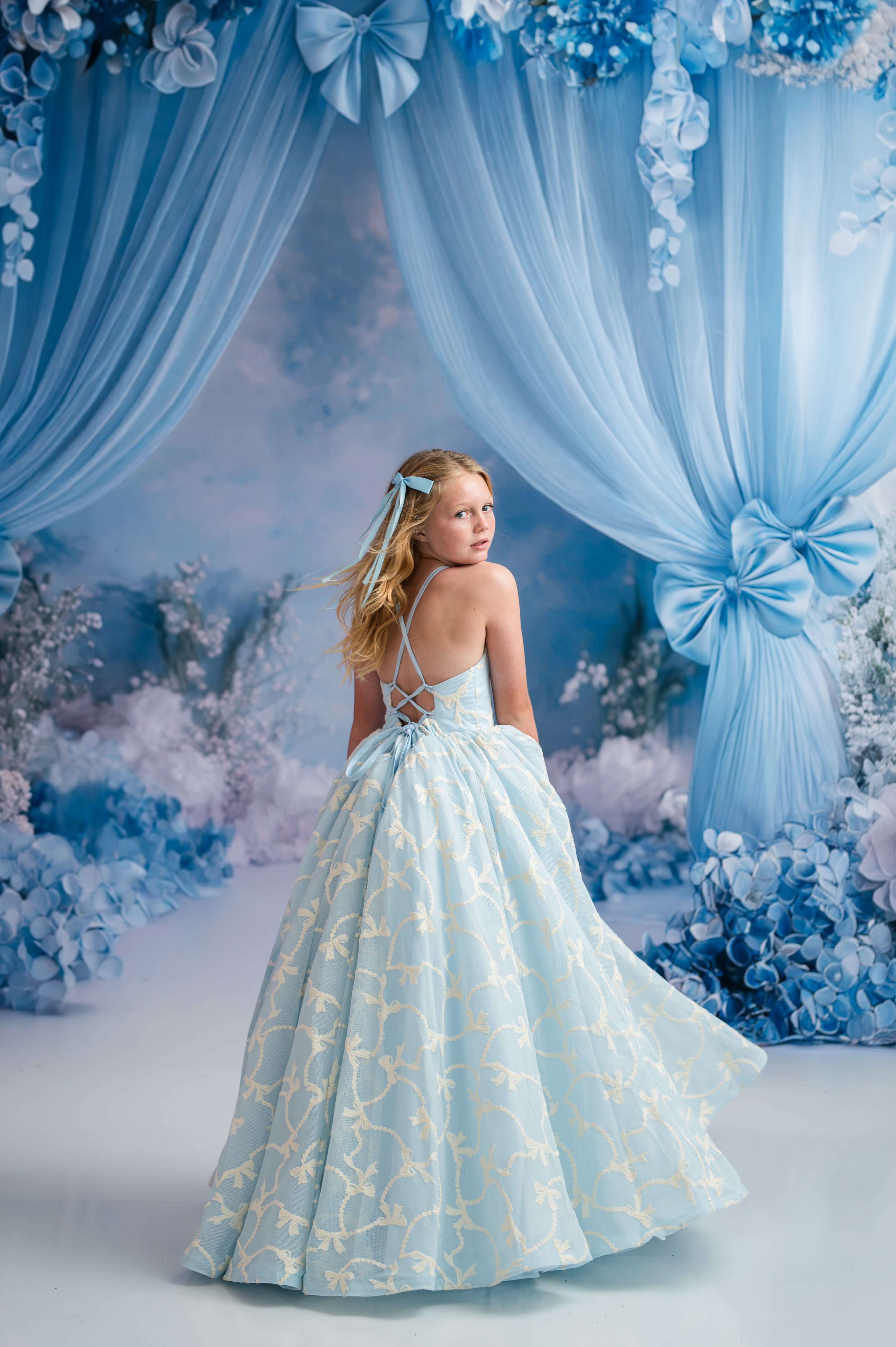 Young girl in a light blue floral dress standing in front of decorative blue curtains and floral arrangements.