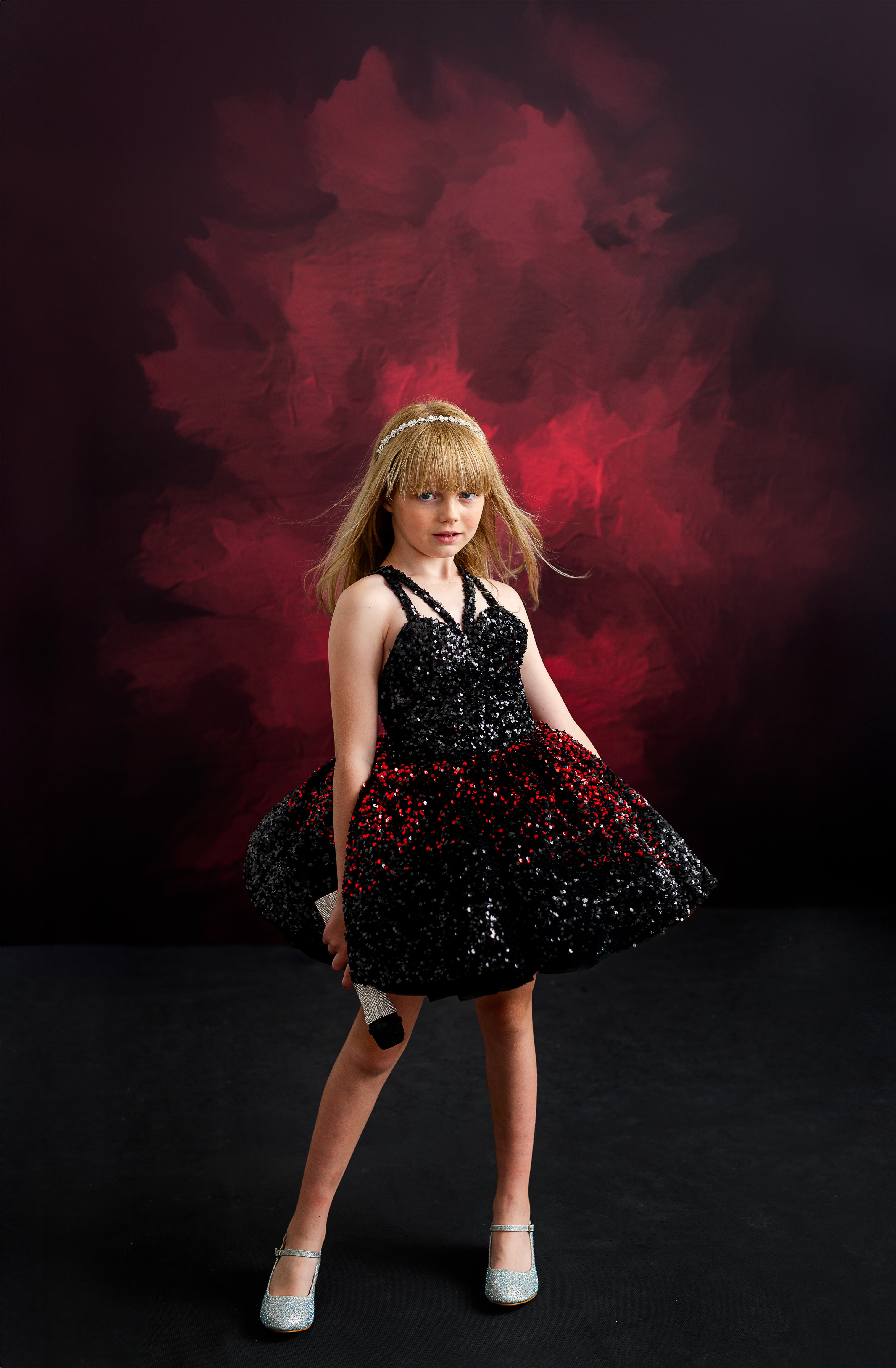 Little girl wearing a dazzling, sequined gown, striking a fierce pose with a crown, capturing Taylor Swift's bold red carpet moments.