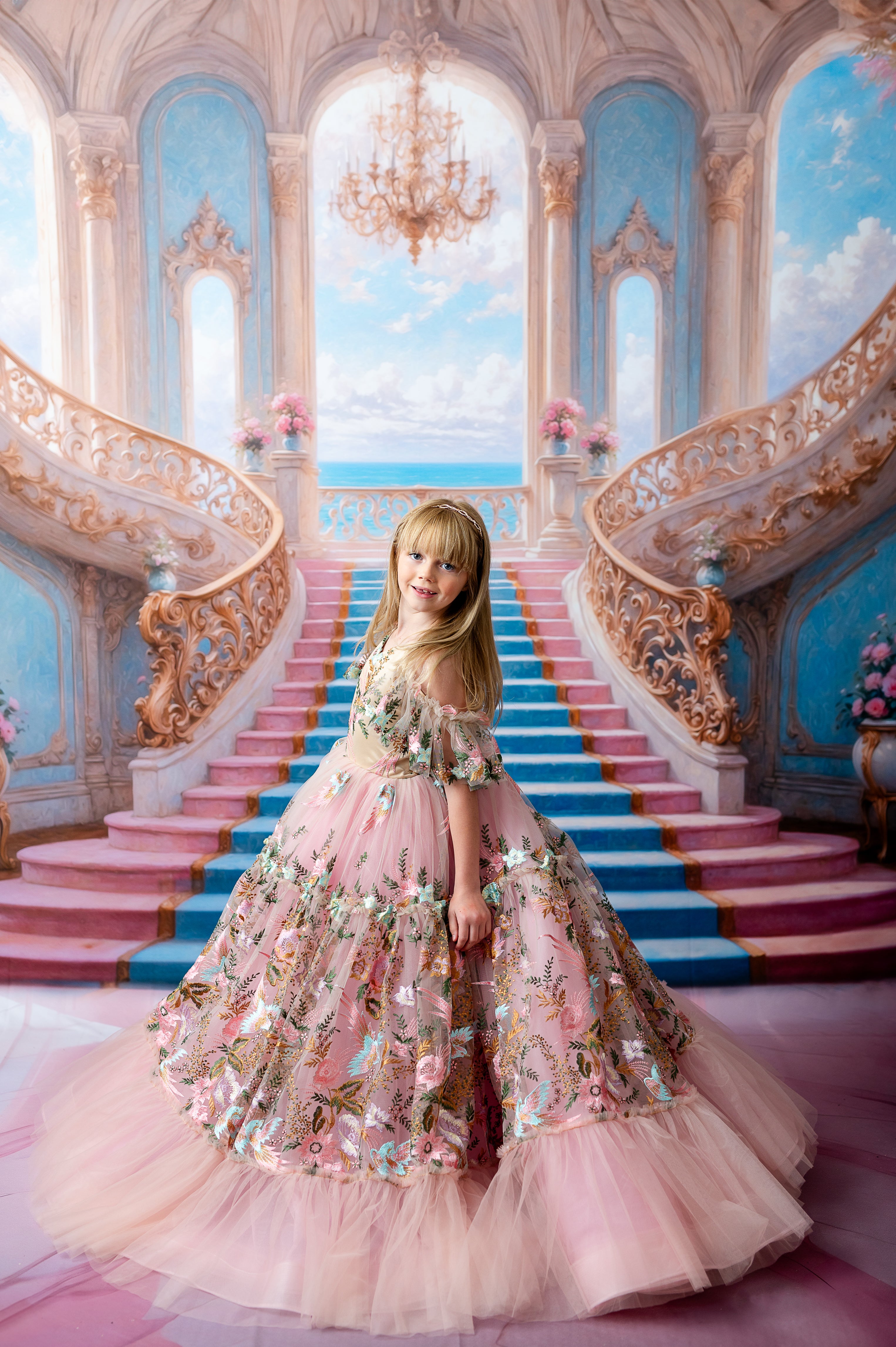 Young girl in a pink floral gown posing in front of a grand staircase with ornate details and a beautiful sky-blue backdrop.