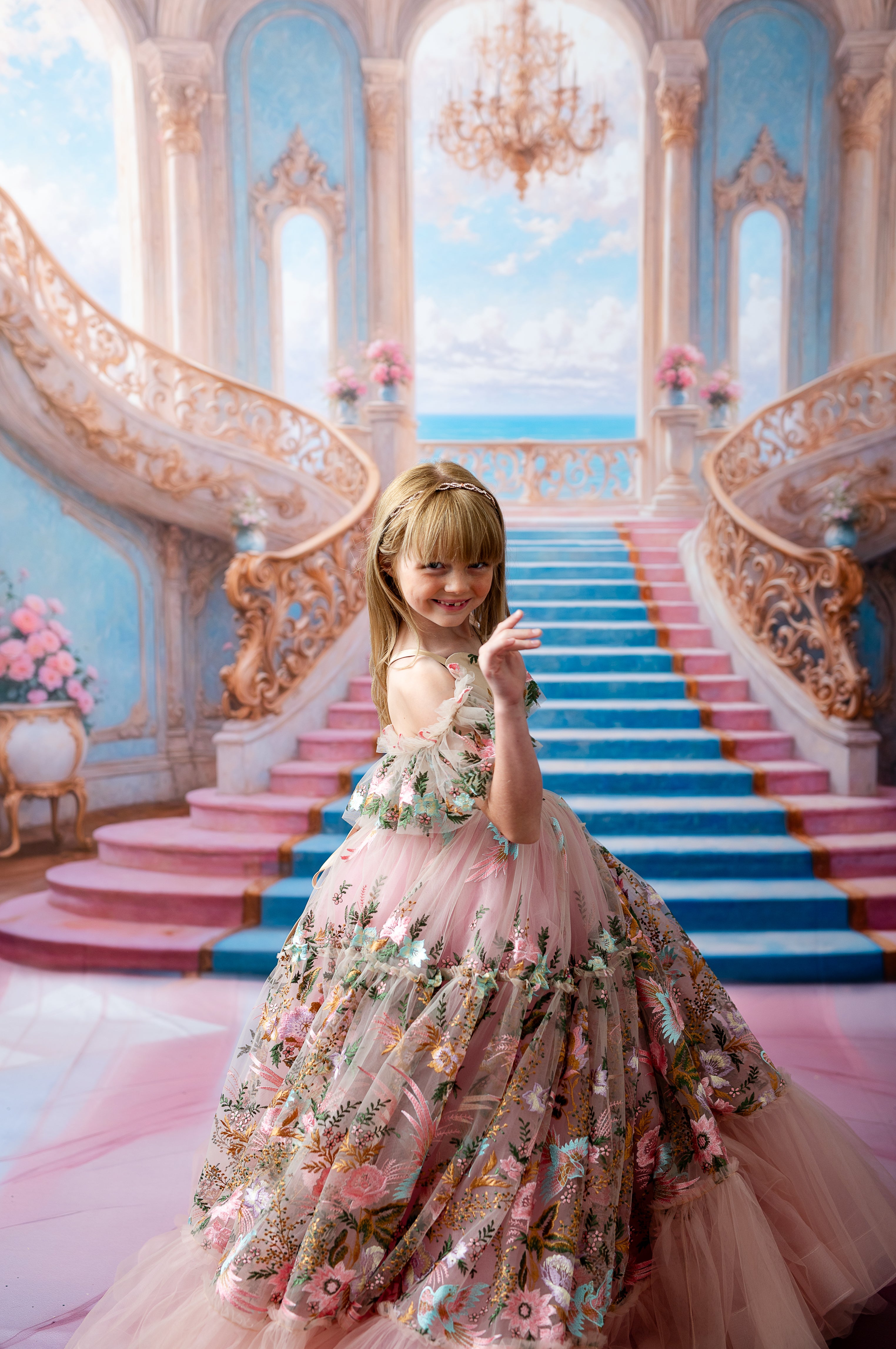 Child wearing a floral embroidered dress with flowing layers of pink tulle, smiling in a luxurious hall with sweeping staircases.