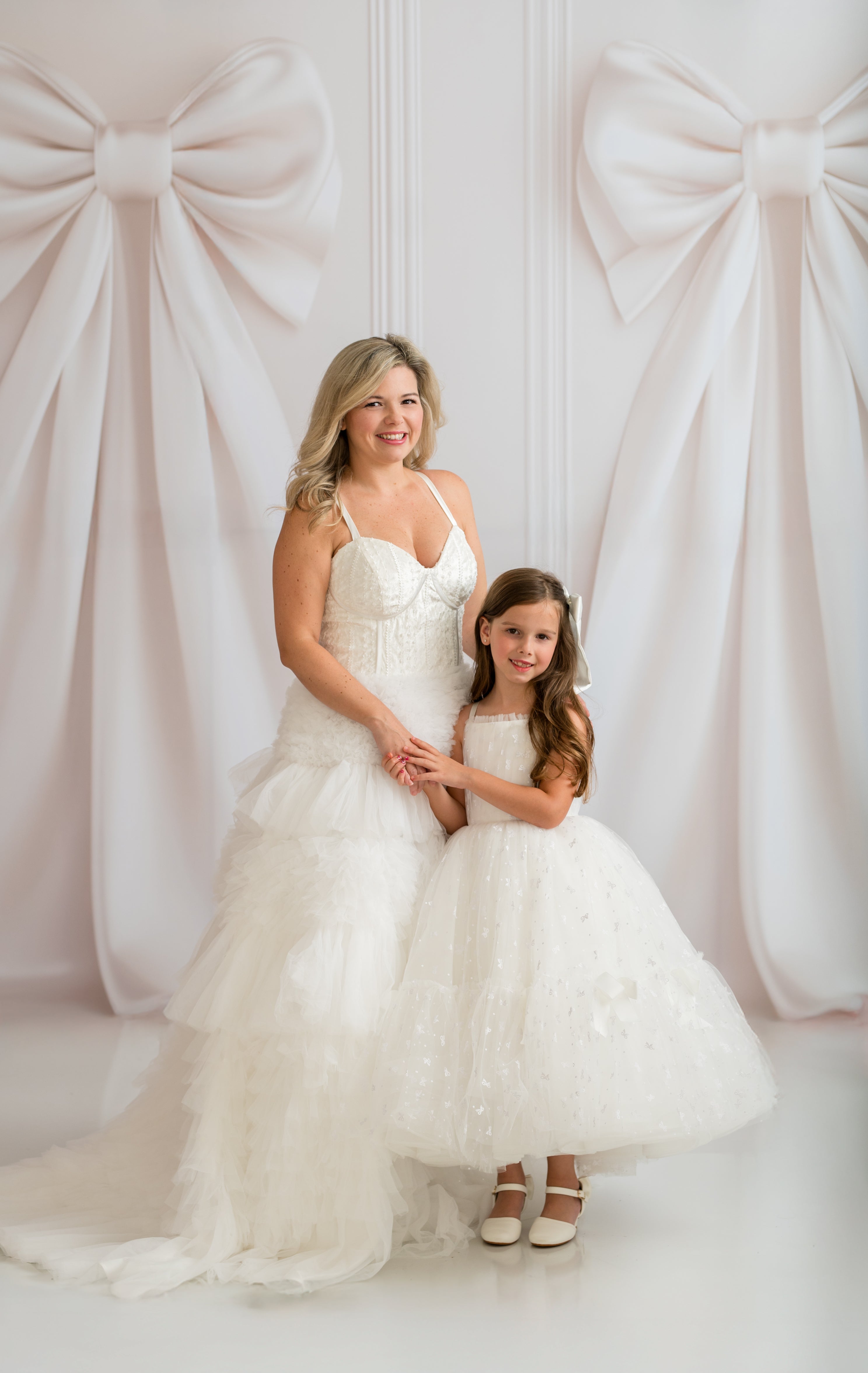 Woman and young girl in white dresses standing in front of a white curtain with large bows.