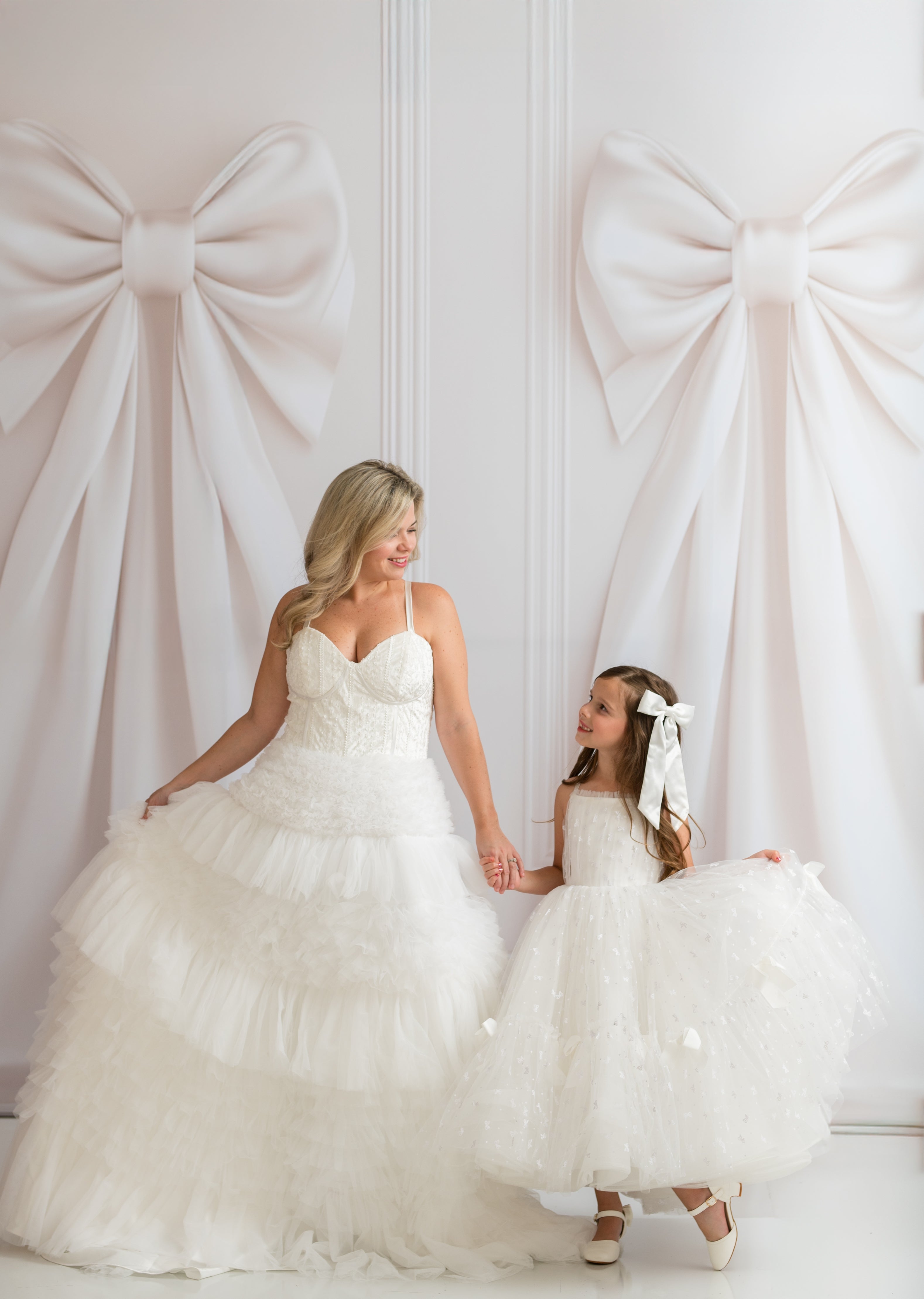 Woman in a white wedding dress holding hands with a young girl in a white dress against a white wall with large decorative bows.