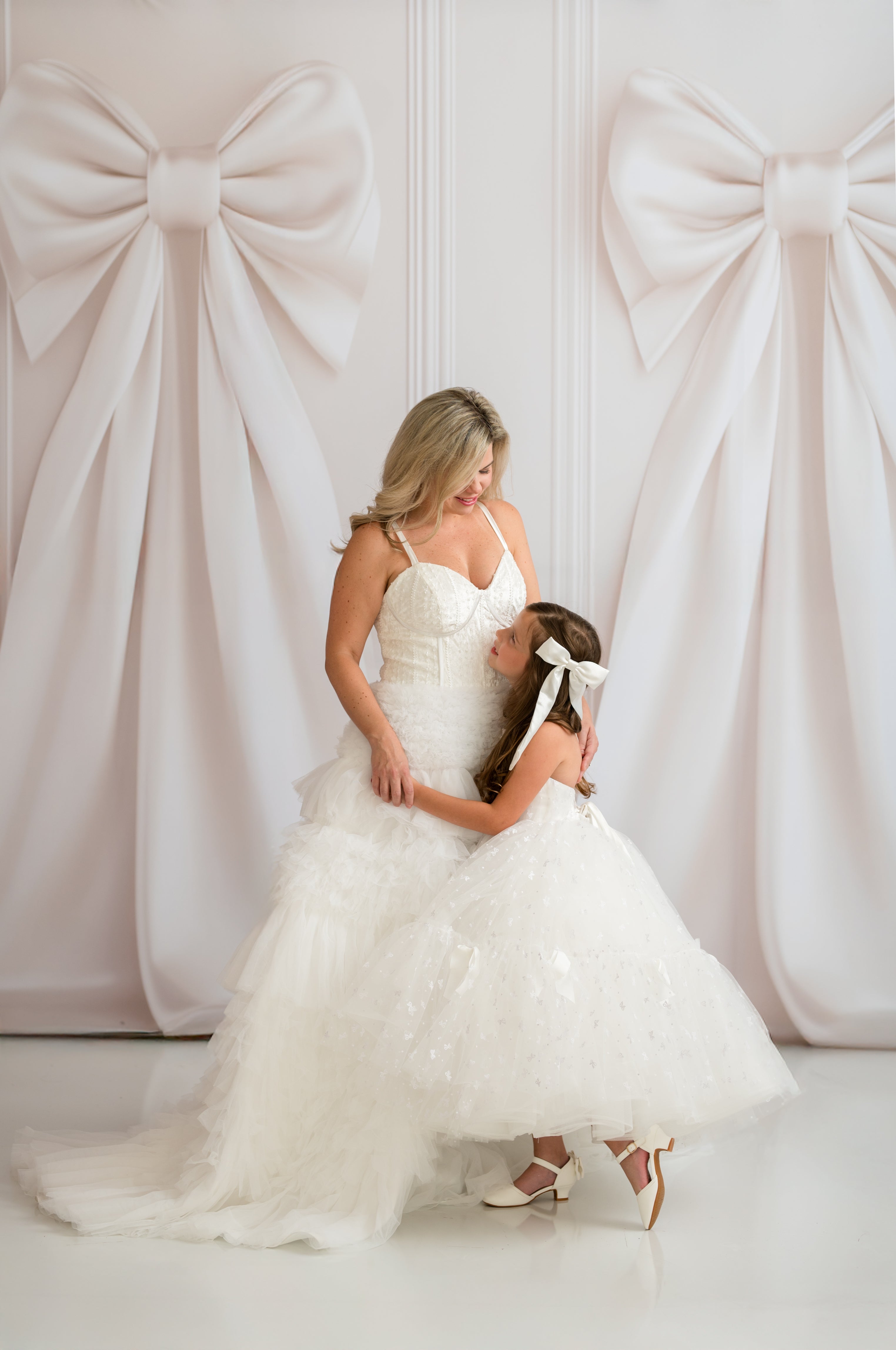 Two women in white dresses standing in front of a decorative wall with large bows.