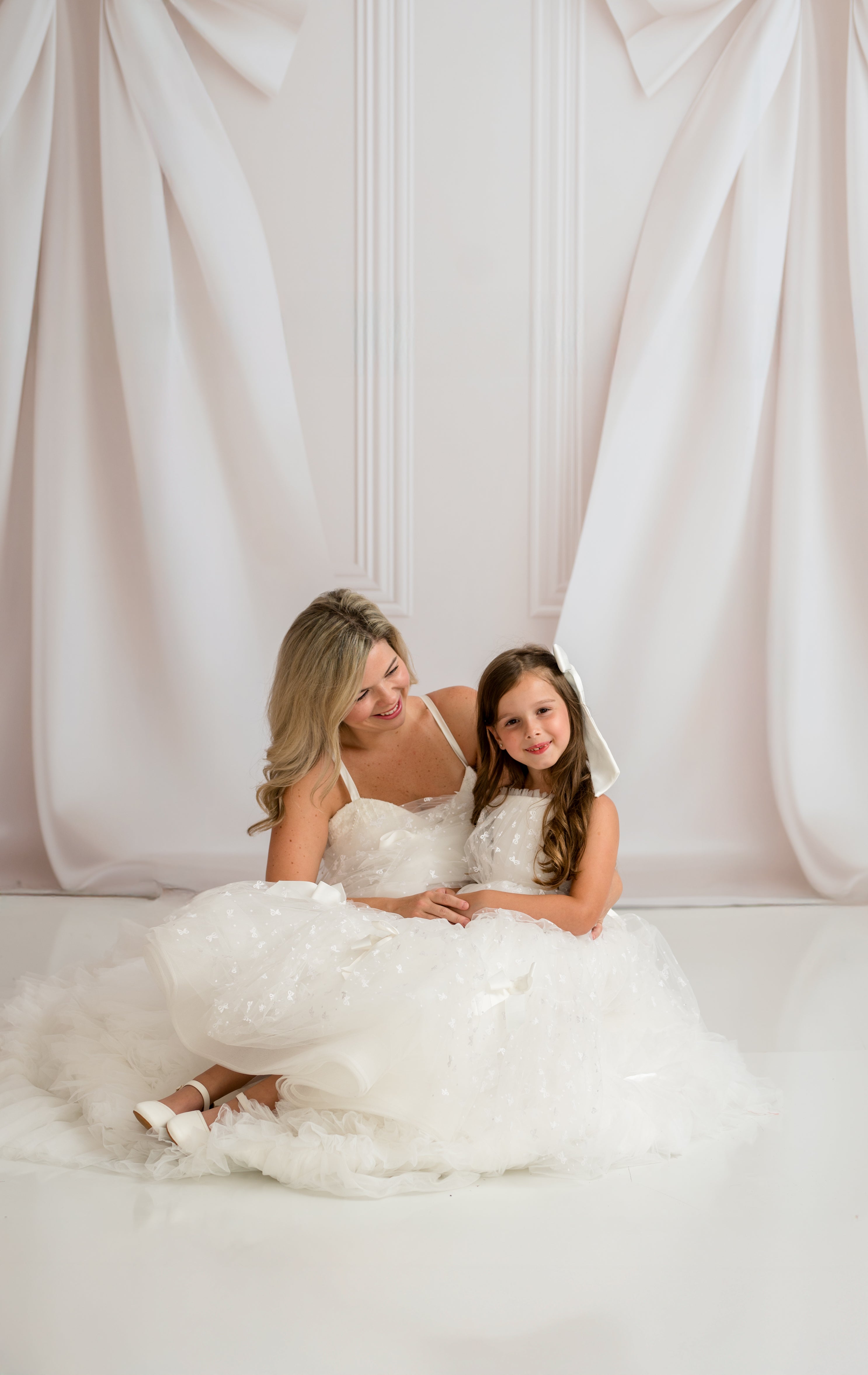 Woman and young girl in white dresses sitting together against a white curtain.