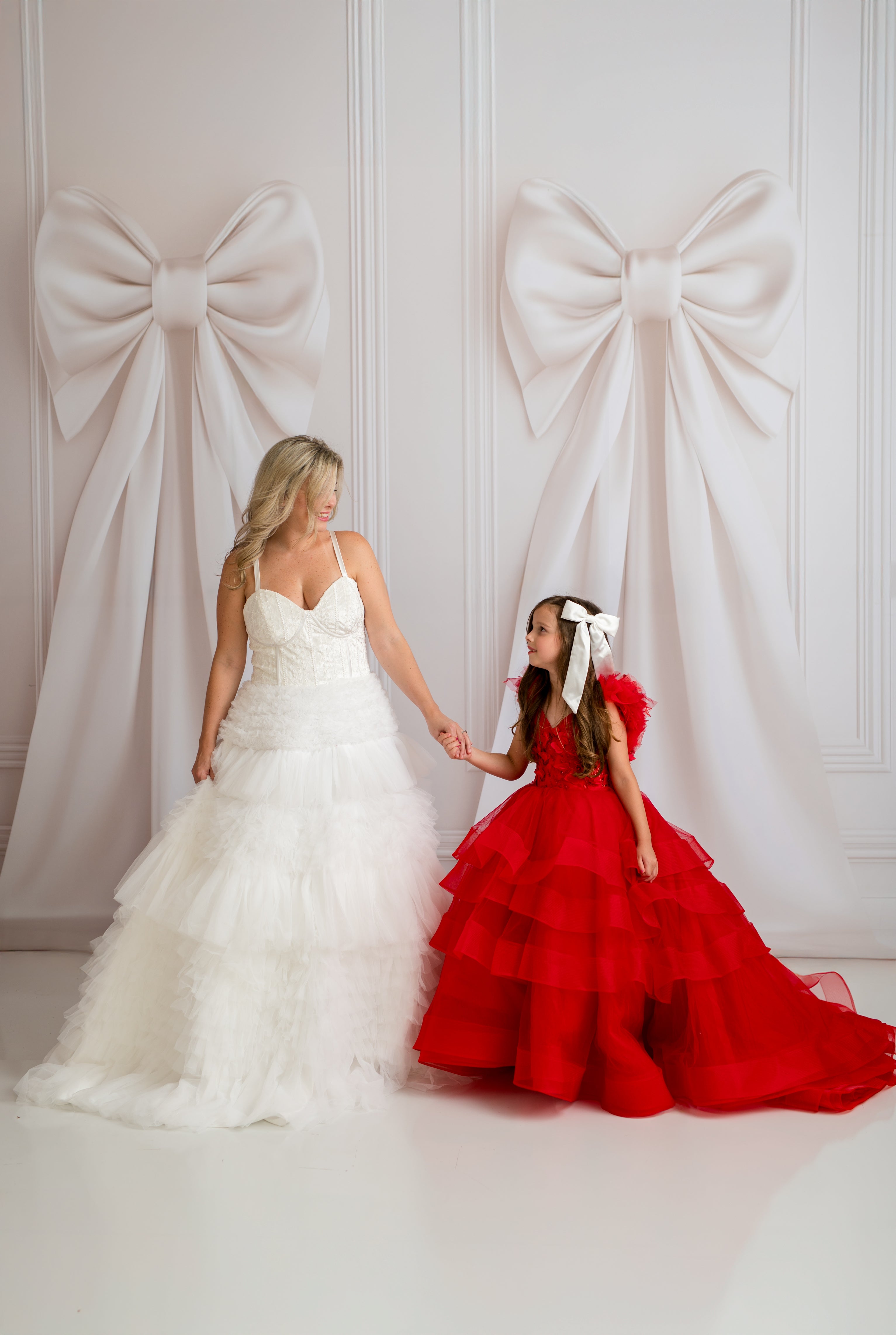Woman in a white dress and young girl in a red dress standing against a white wall with large decorative bows.