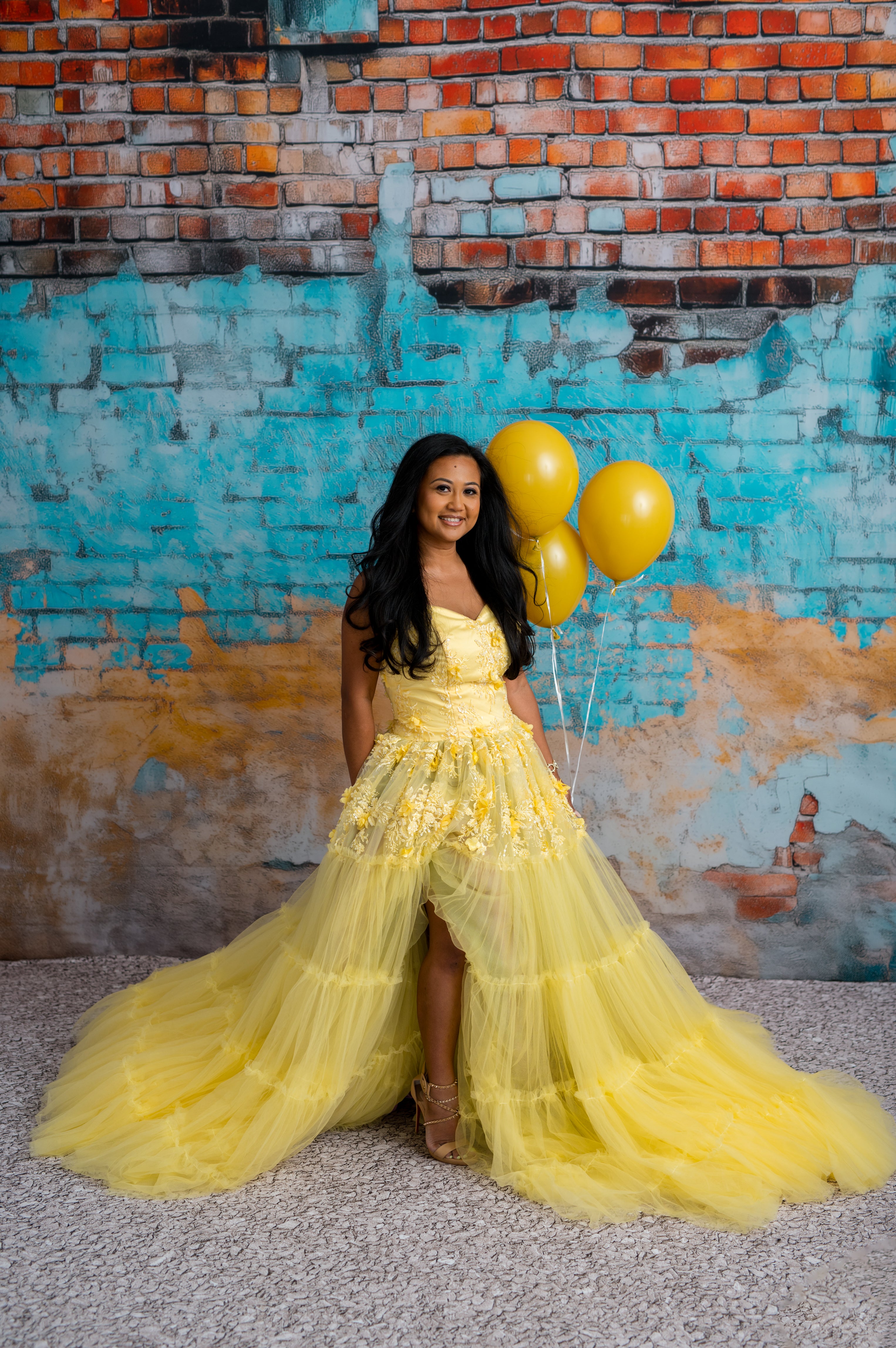 Woman in a yellow dress holding balloons against a colorful brick wall.