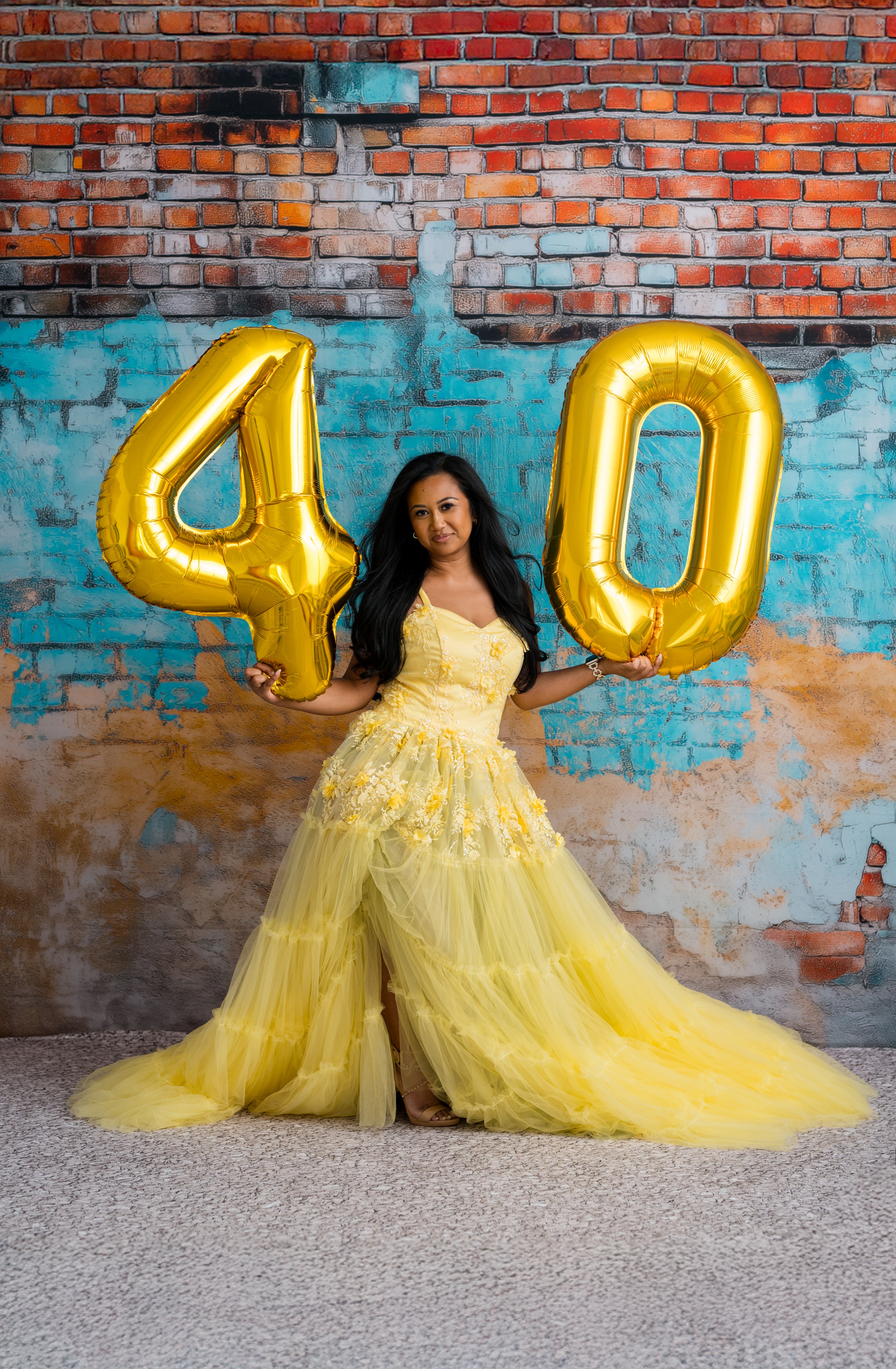 Woman in a yellow dress holding gold number balloons against a colorful brick wall.