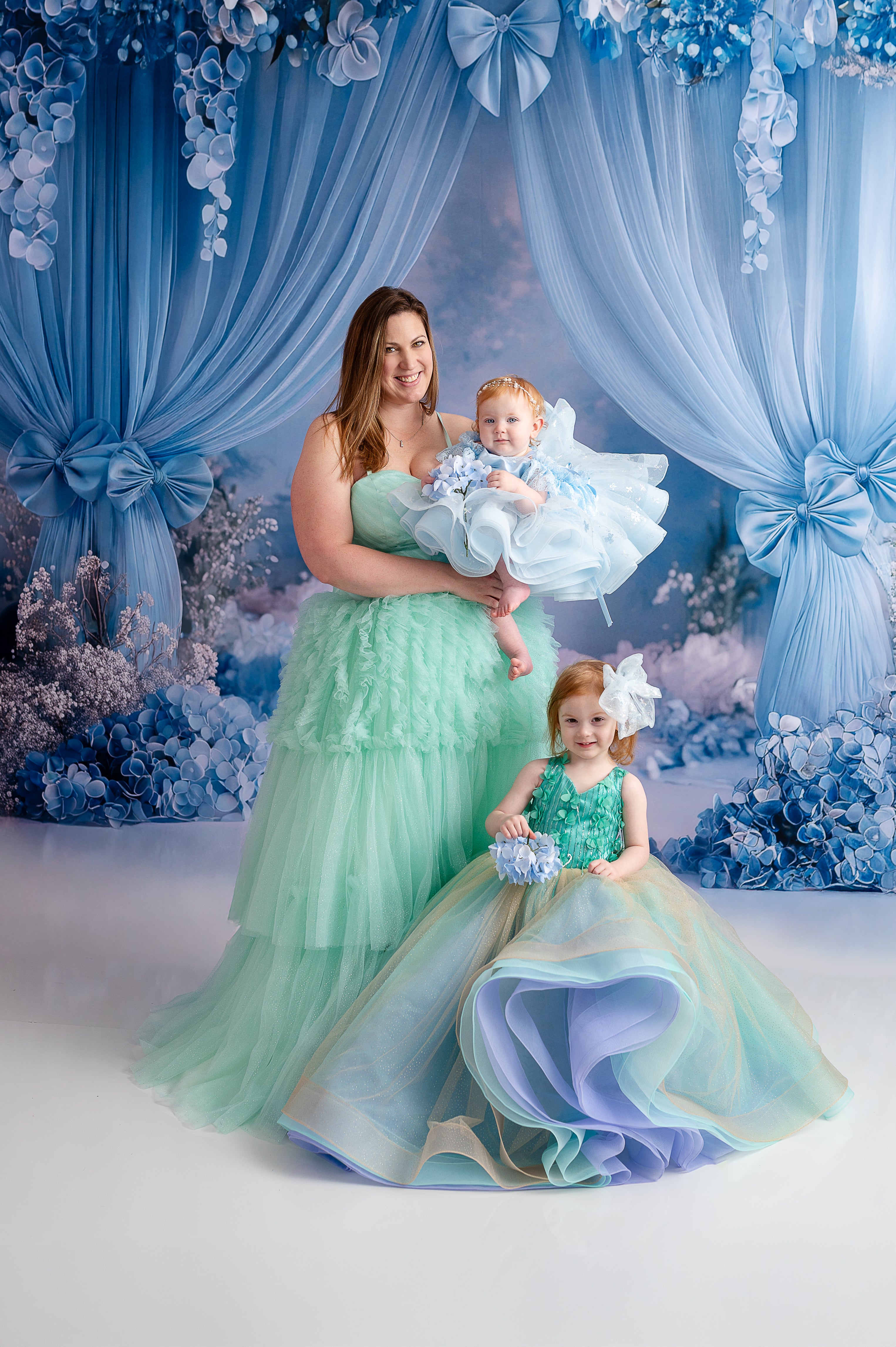 A mother and daughter hold hands, dressed in matching mint green gowns, standing in front of a soft floral backdrop.