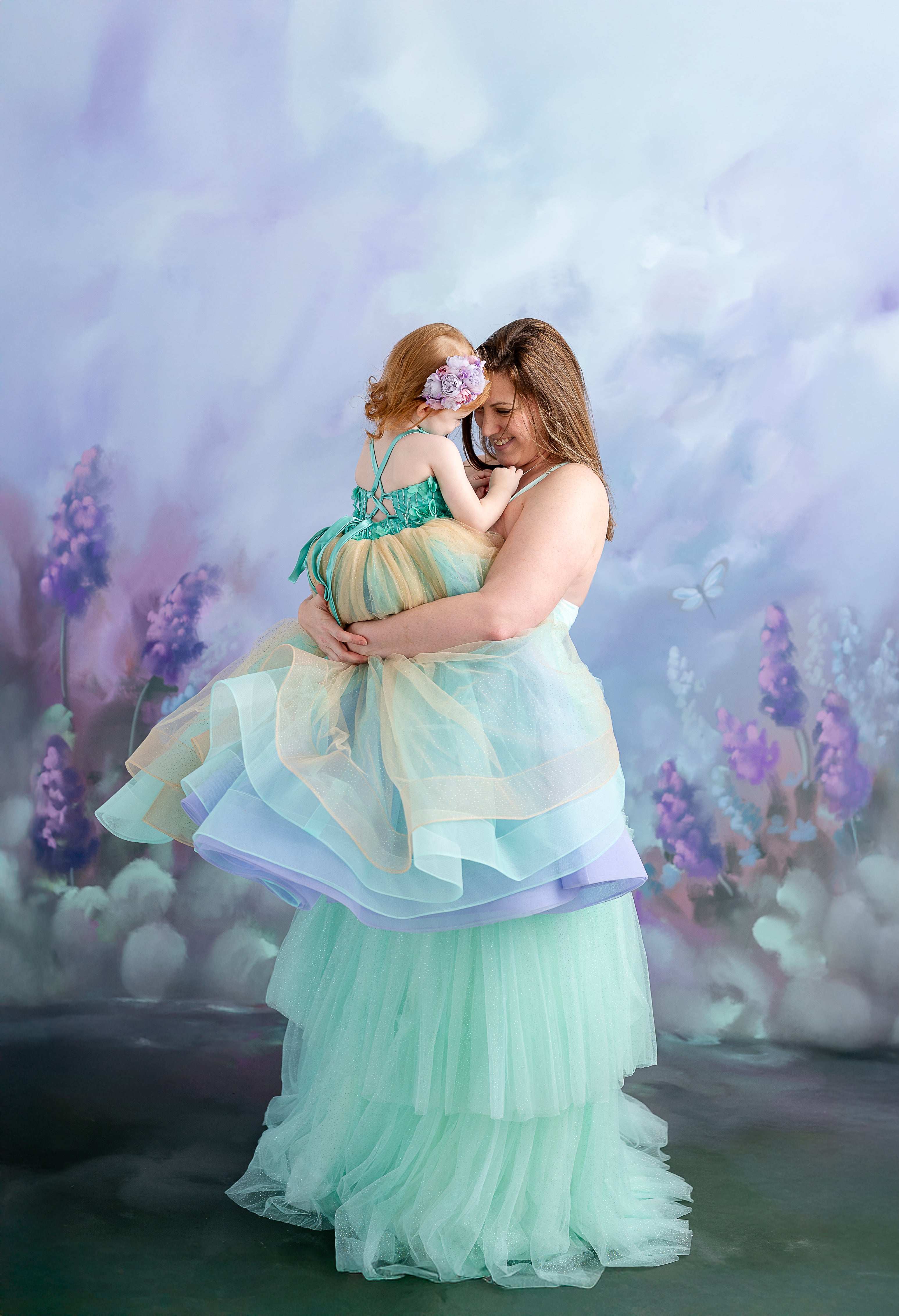 A little girl with a floral hair accessory holds her mother’s hand while dressed in a fairytale-inspired gown.
