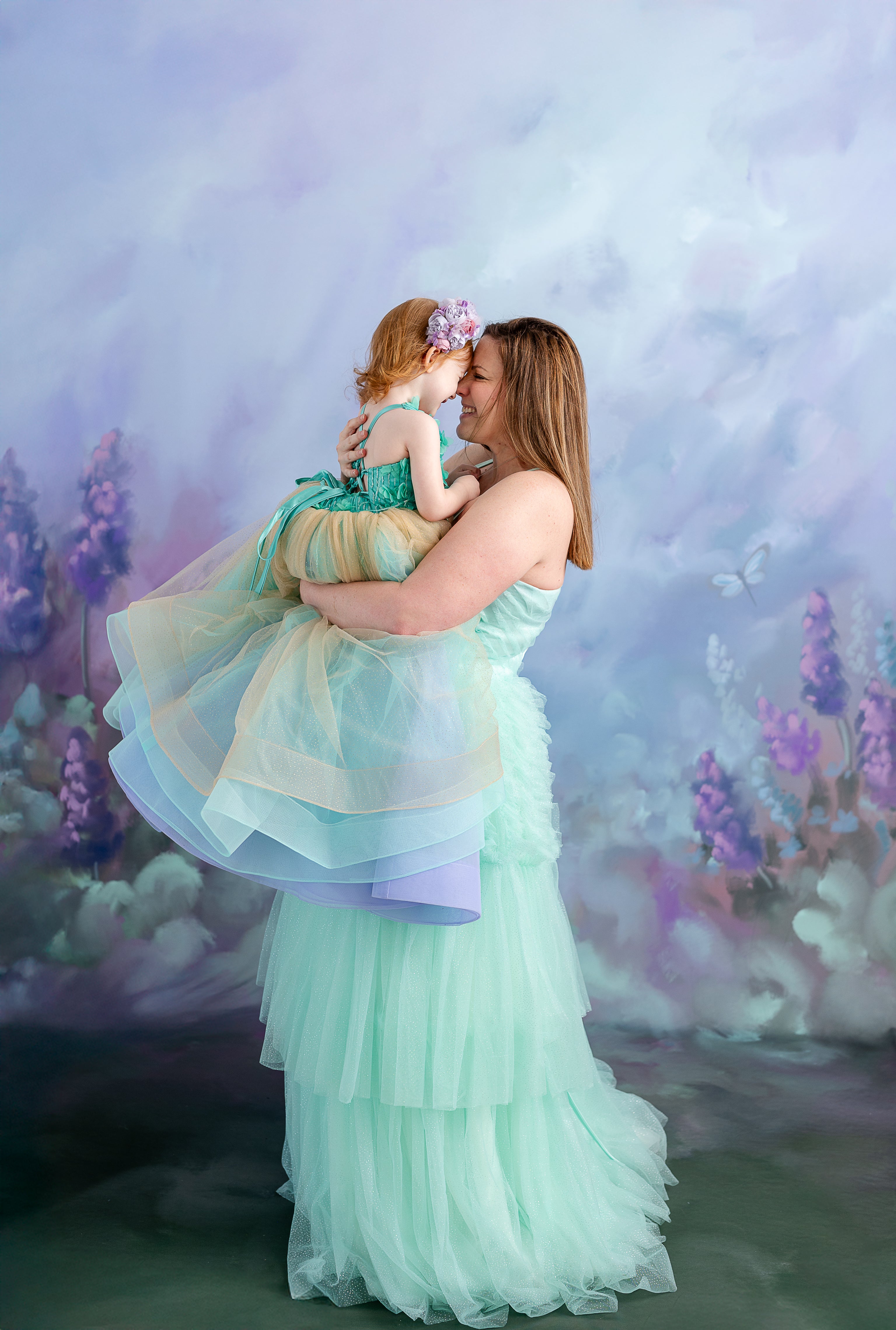 A mother and daughter hold hands, dressed in pastel gowns, standing in front of a dreamy floral backdrop.