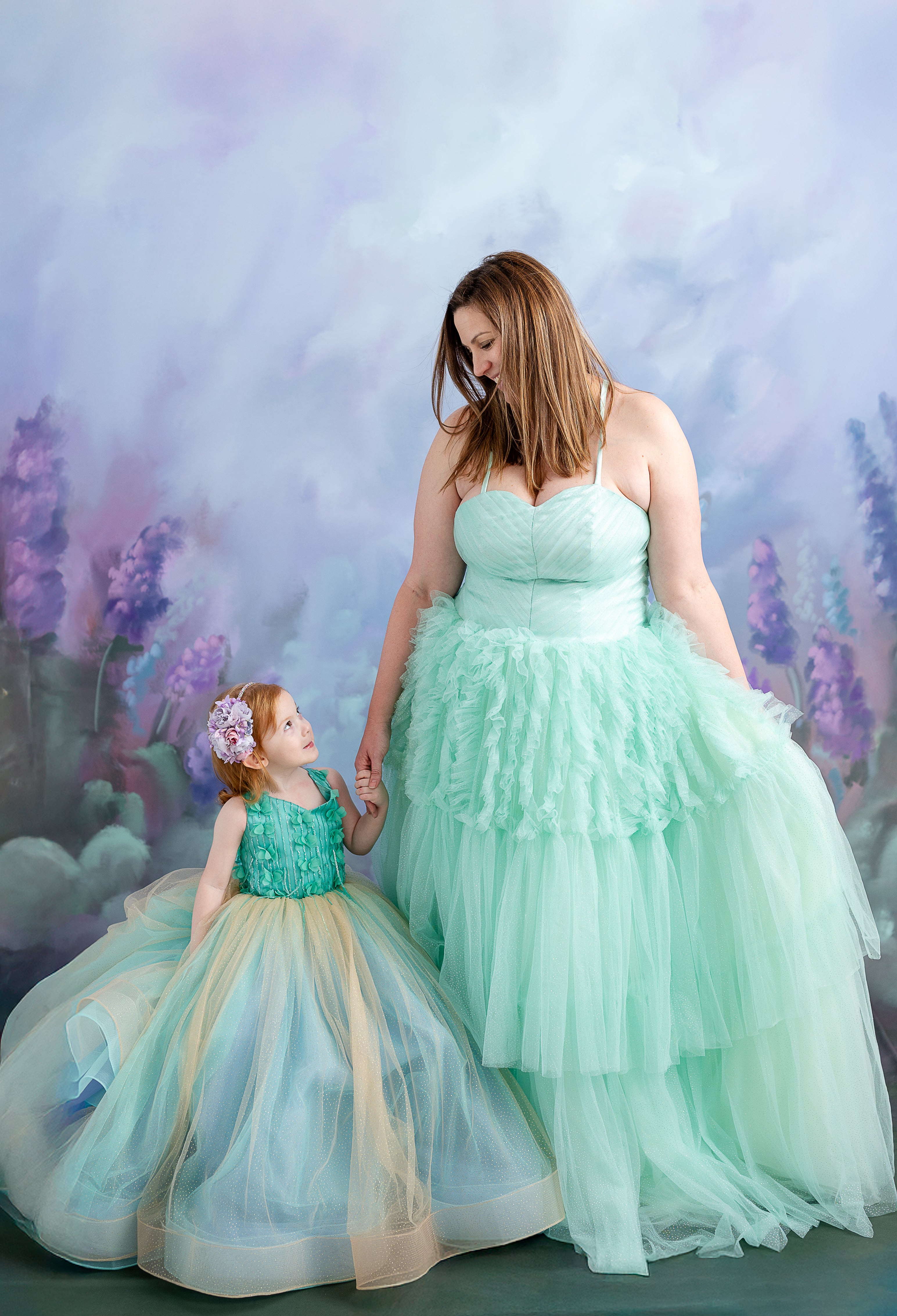 A little girl with red hair and a flower headpiece gazes up at her mother, both wearing elegant dresses.