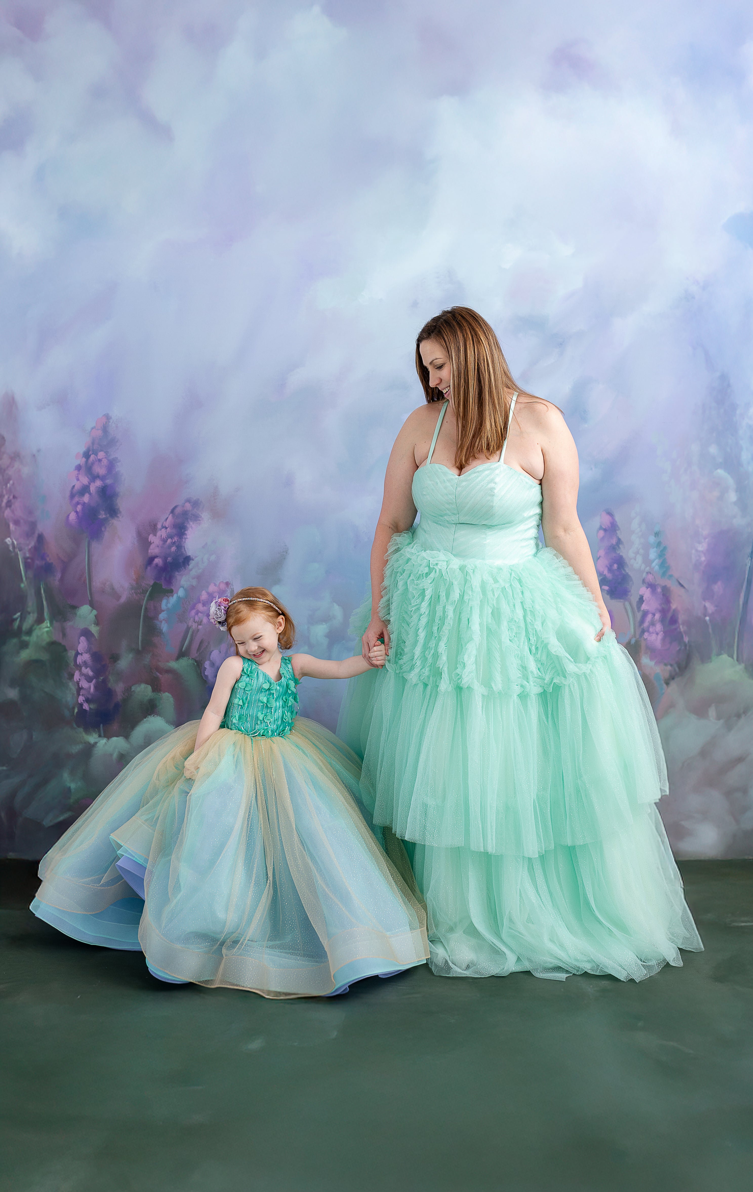 A woman and child, dressed in coordinating pastel gowns, share a tender moment in front of a soft, painted backdrop.