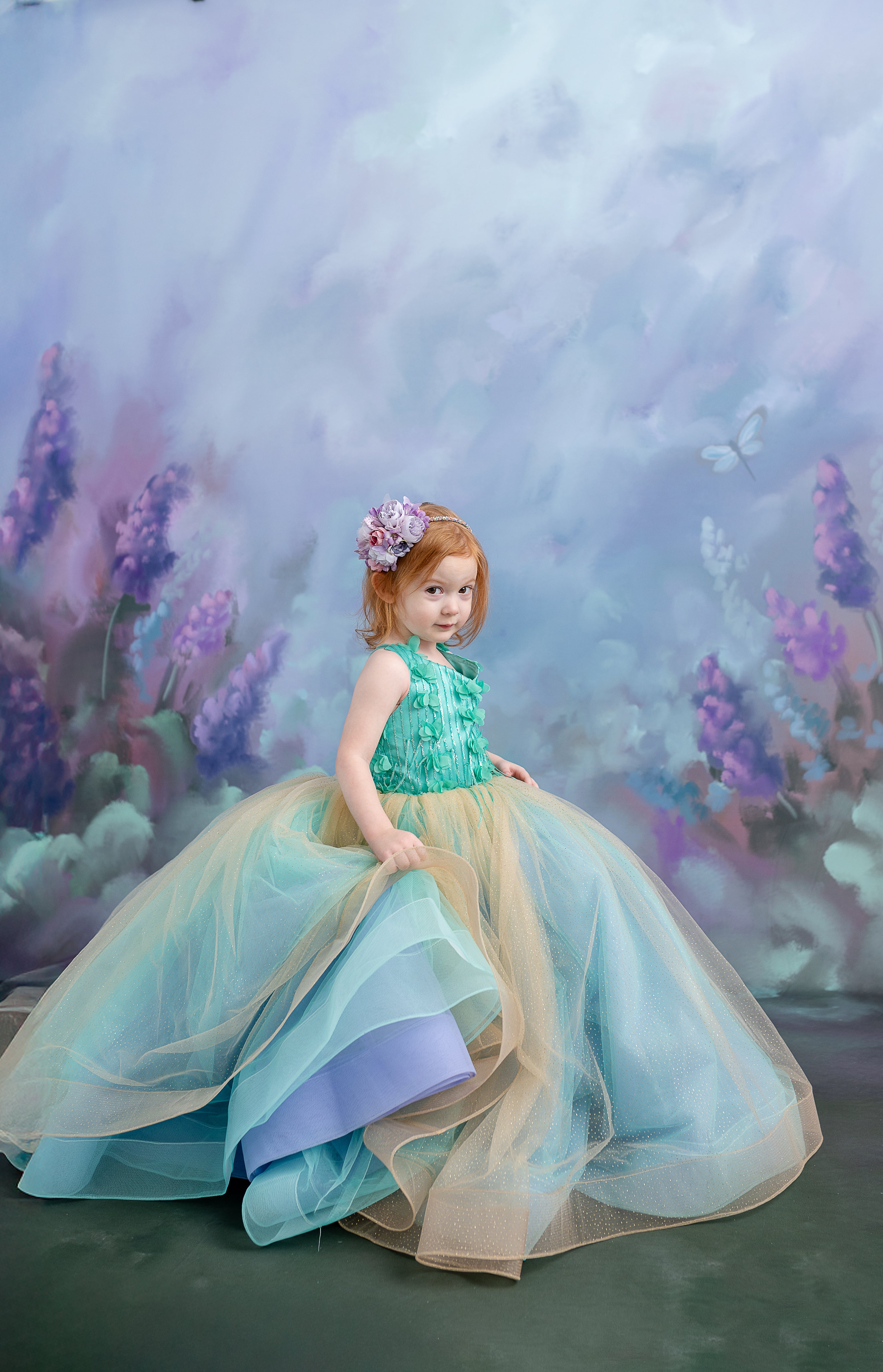 Toddler in the Evelyn gown posing with a dreamy expression, surrounded by blue floral elements.