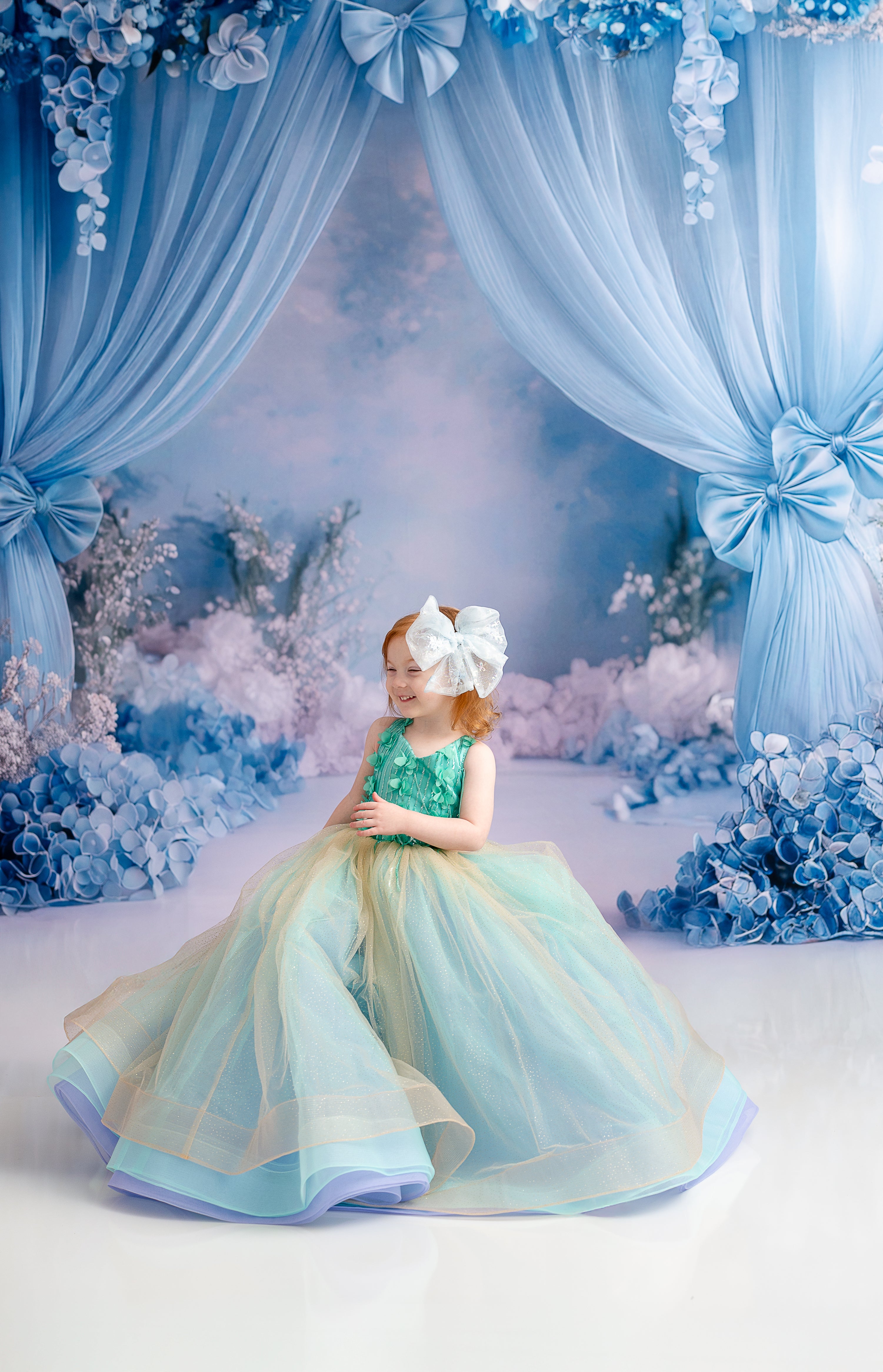 A little girl smiling while holding a bouquet of blue flowers, wearing the Evelyn gown in an elegant setting.