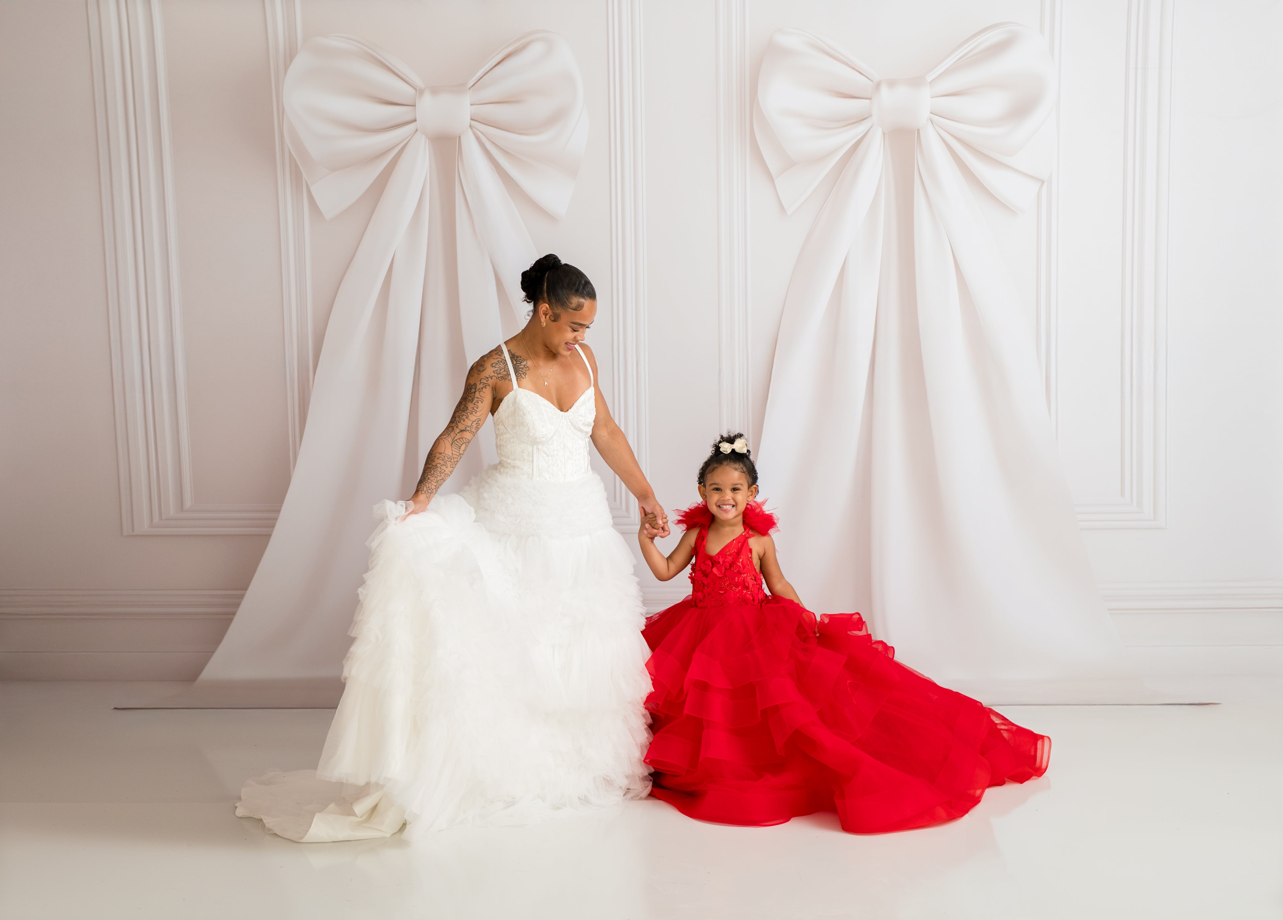 Woman in a white wedding dress holding hands with a young girl in a red dress against a decorative white wall.