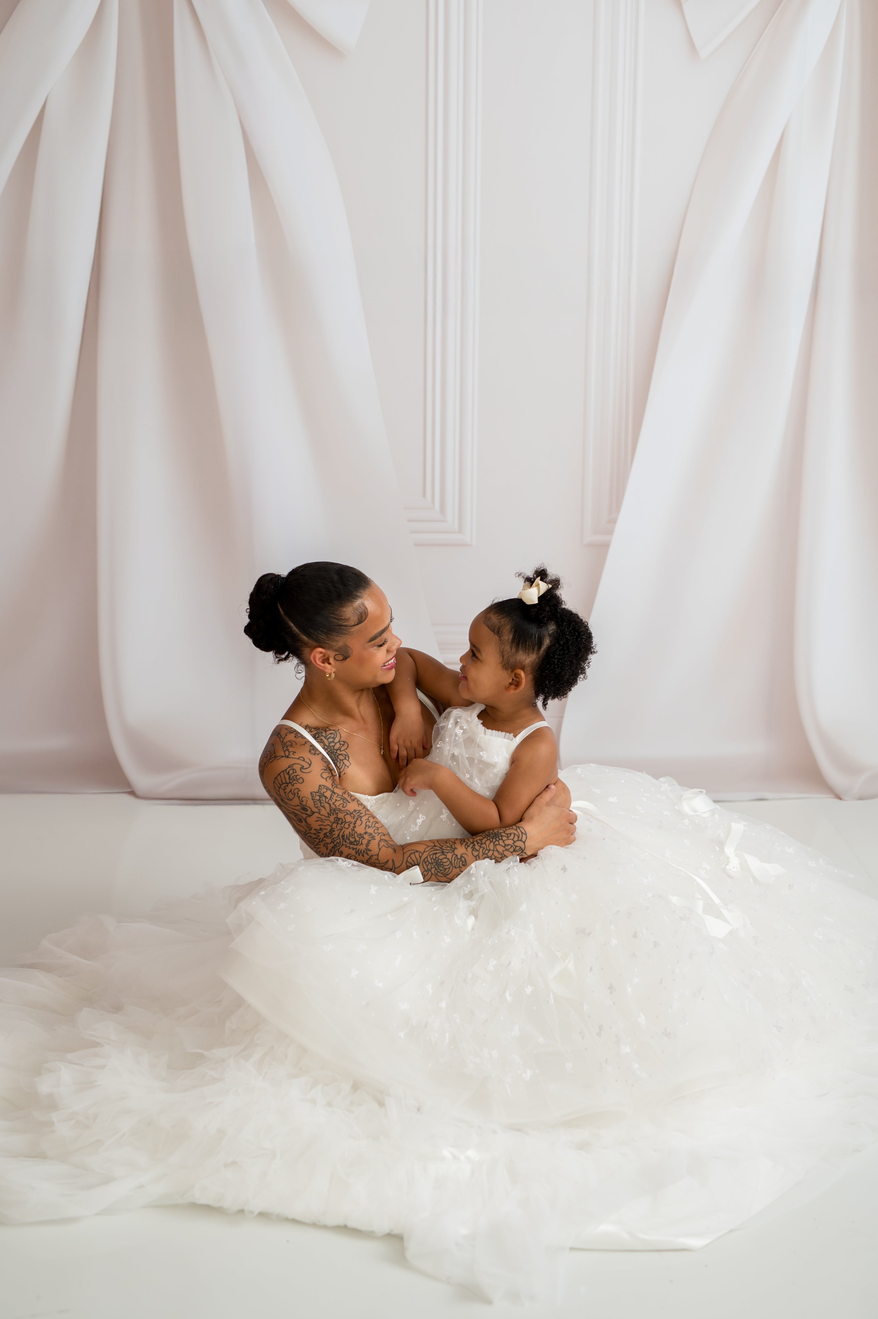 Woman and child in white dresses sitting together against a white curtain.
