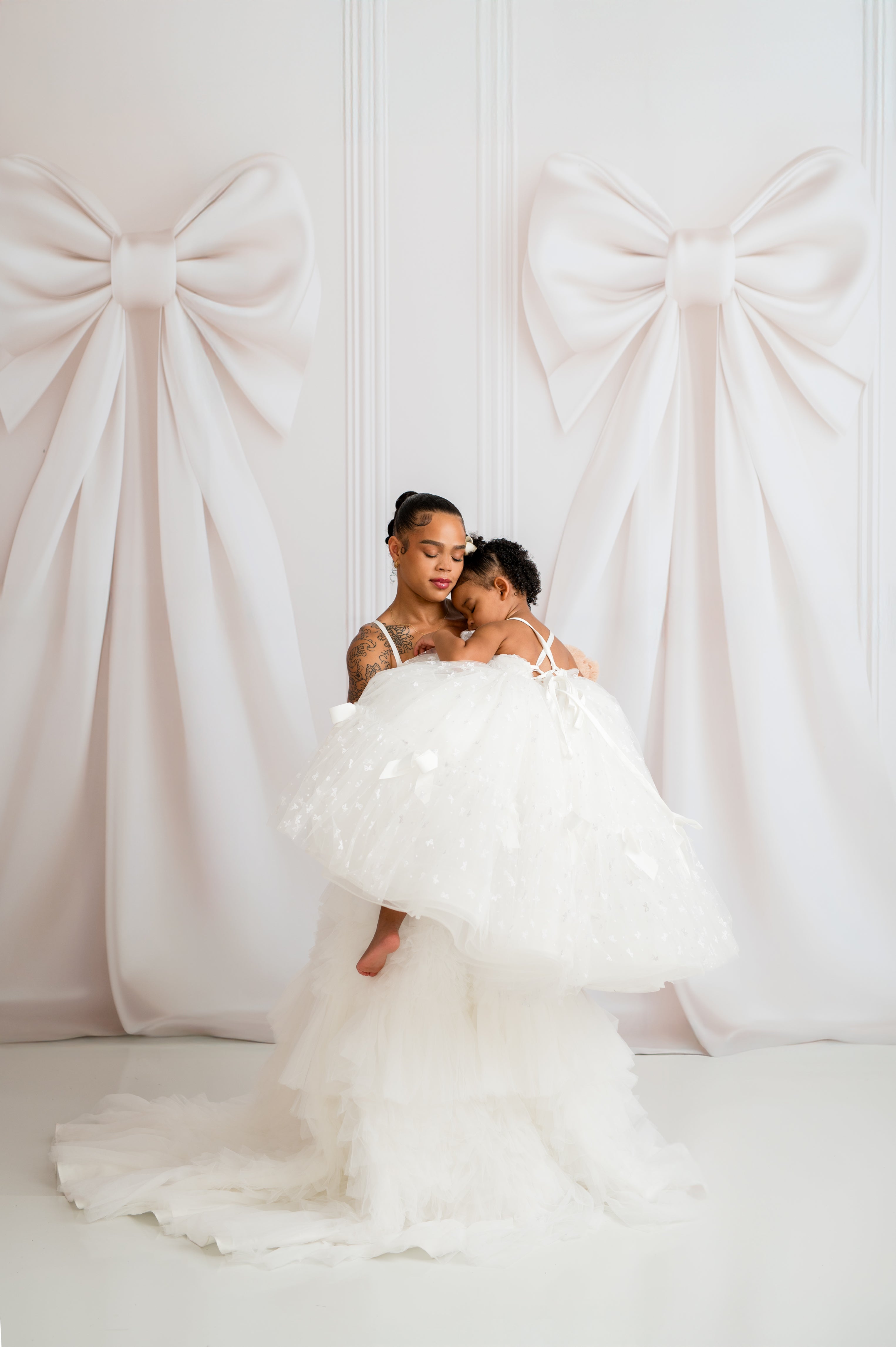 Two women in white dresses standing in front of a decorative white wall with large bows.