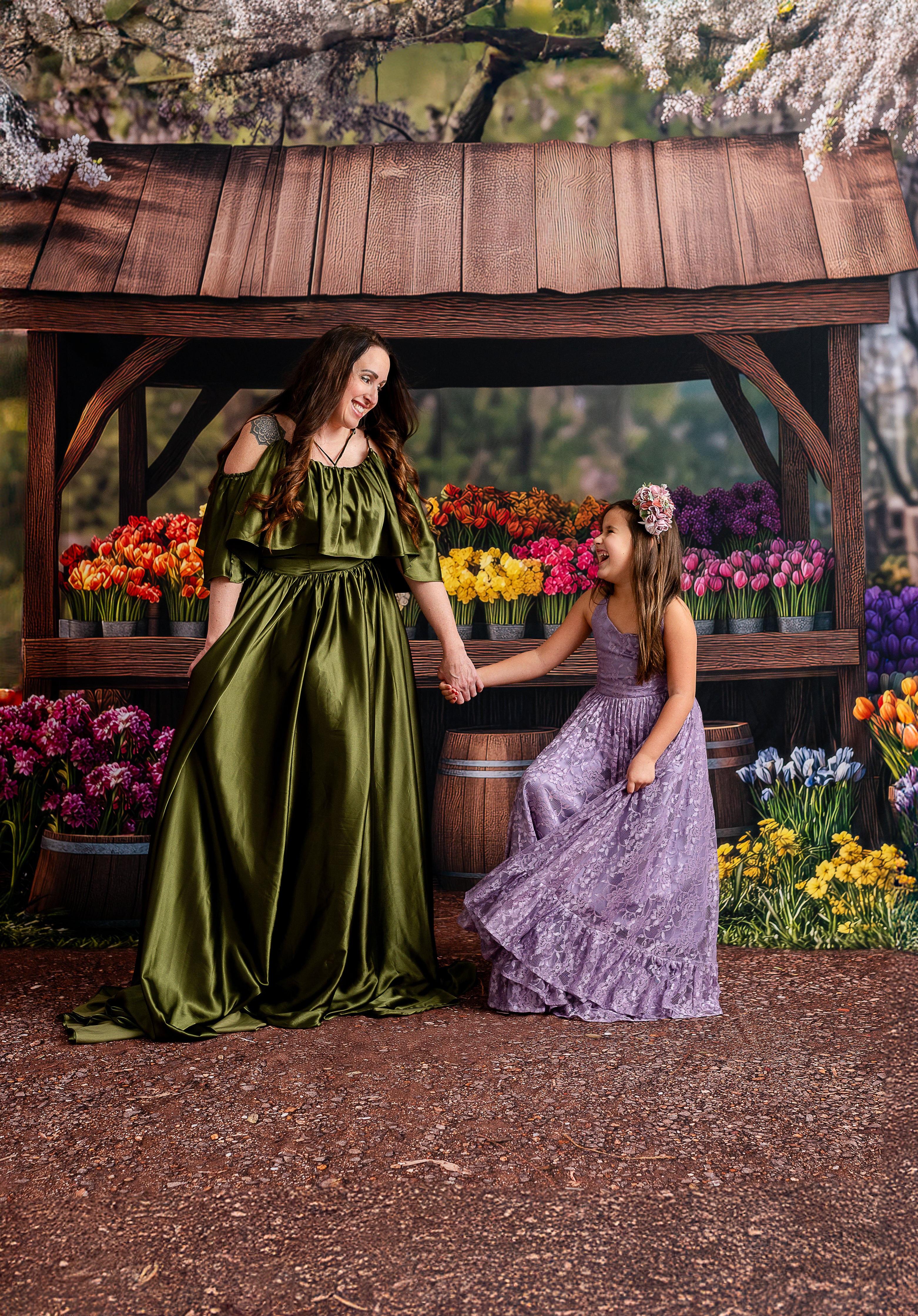 "A mother and daughter holding hands in front of a rustic flower market backdrop, wearing elegant green and lavender gowns."