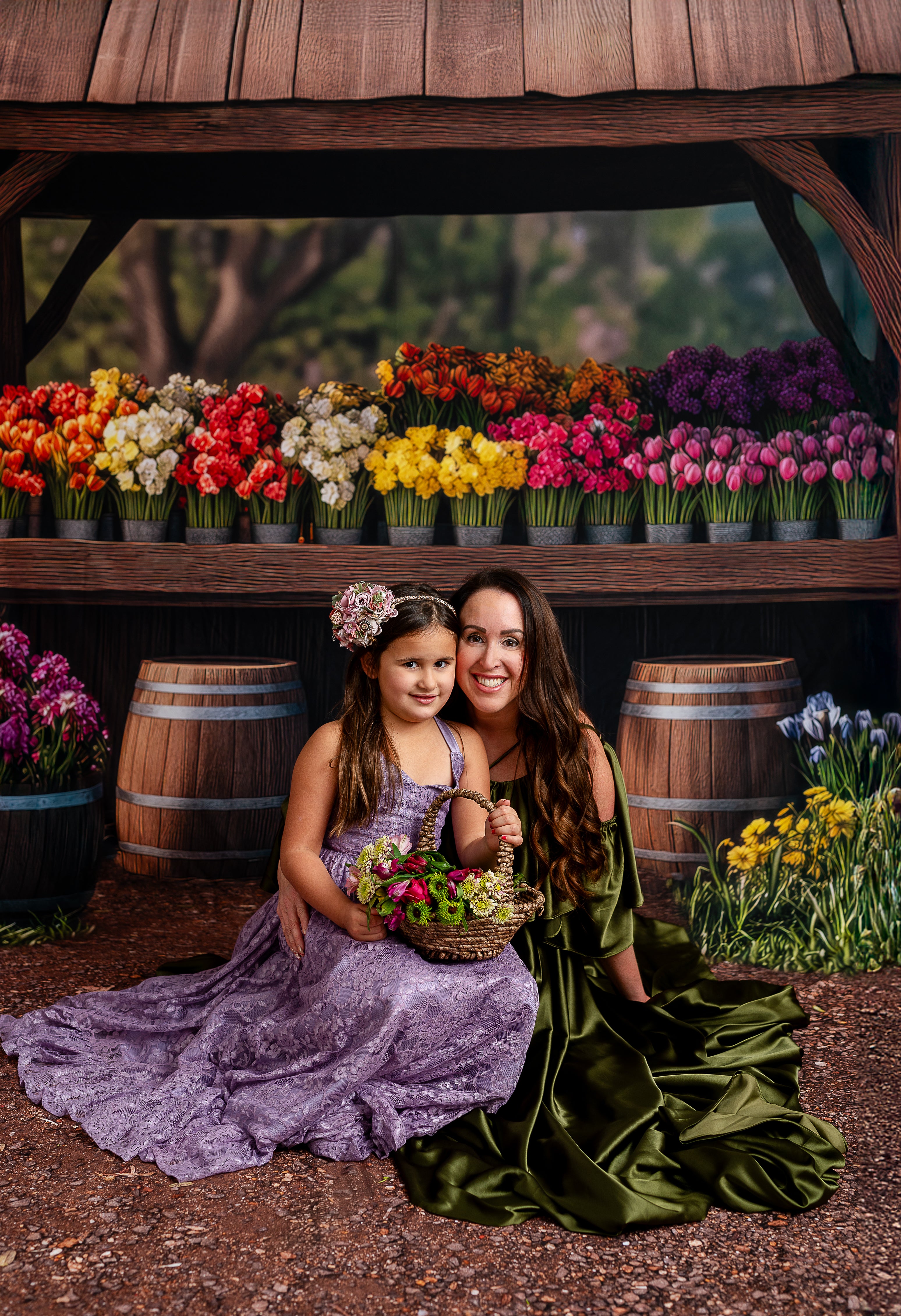 "A woman in a green satin gown smiles at a young girl in a purple lace dress, surrounded by vibrant flowers in barrels."