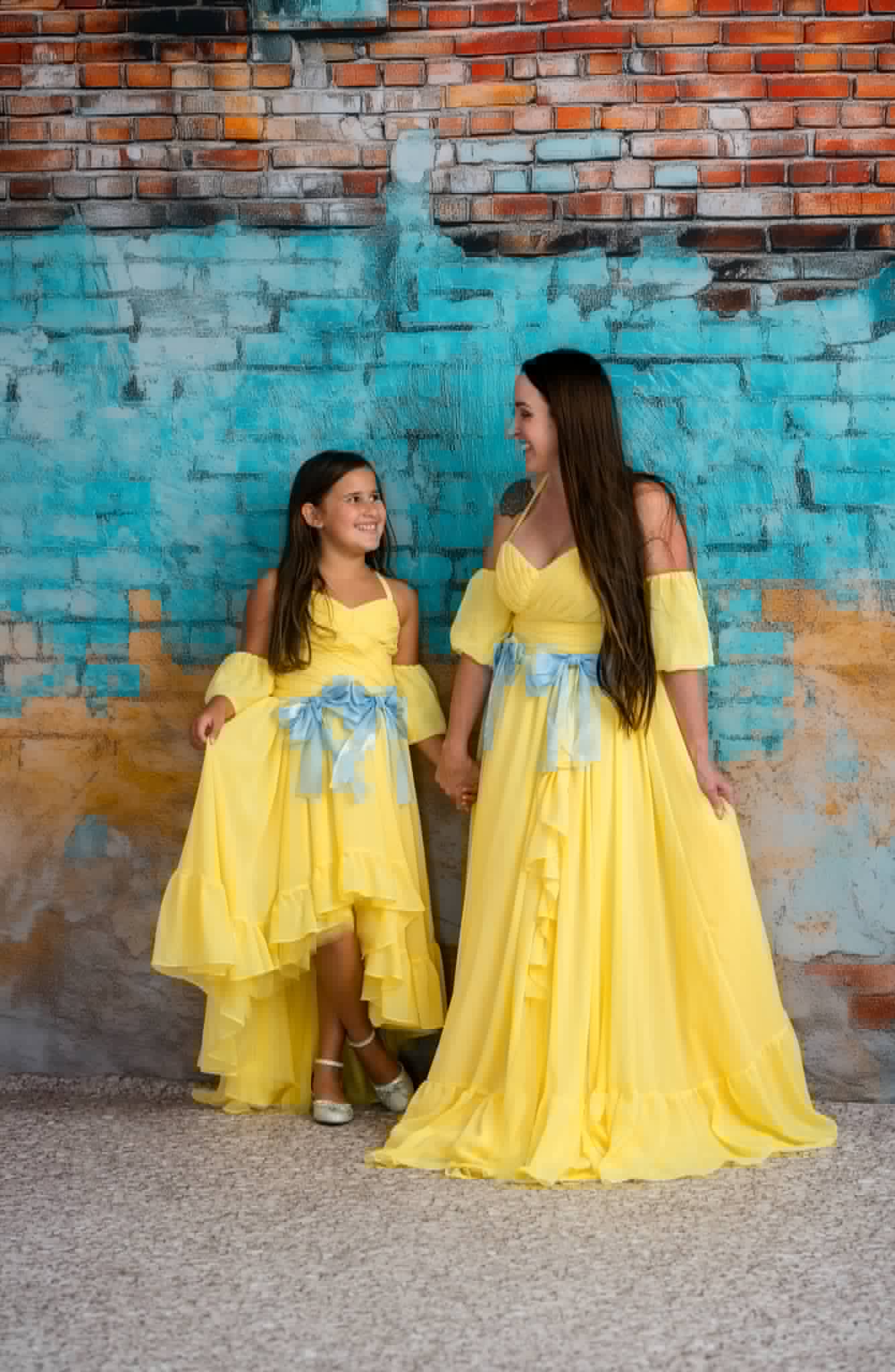 Two women in yellow dresses standing against a textured wall.