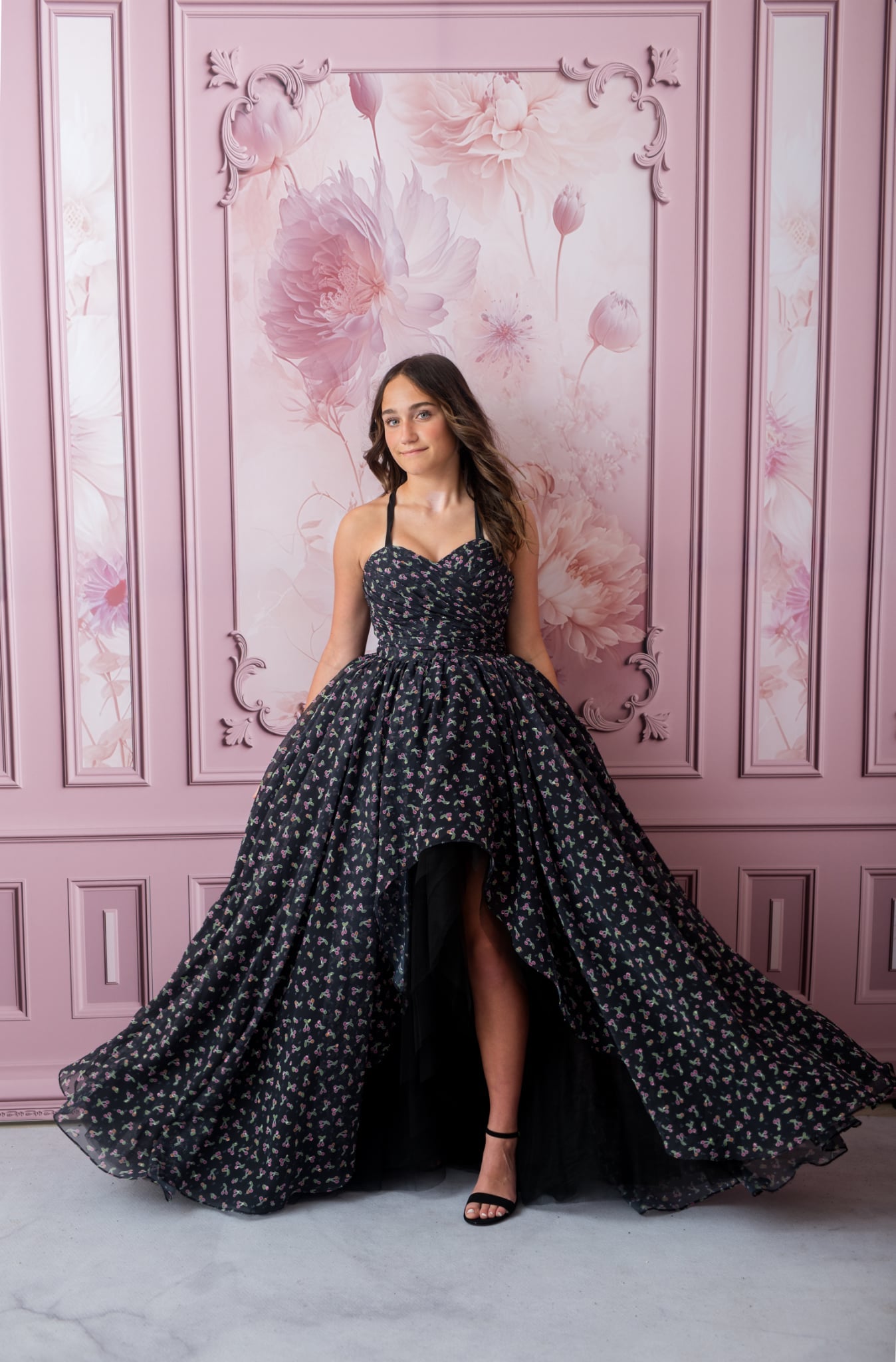 Woman in a black floral dress standing against a pink floral wall.