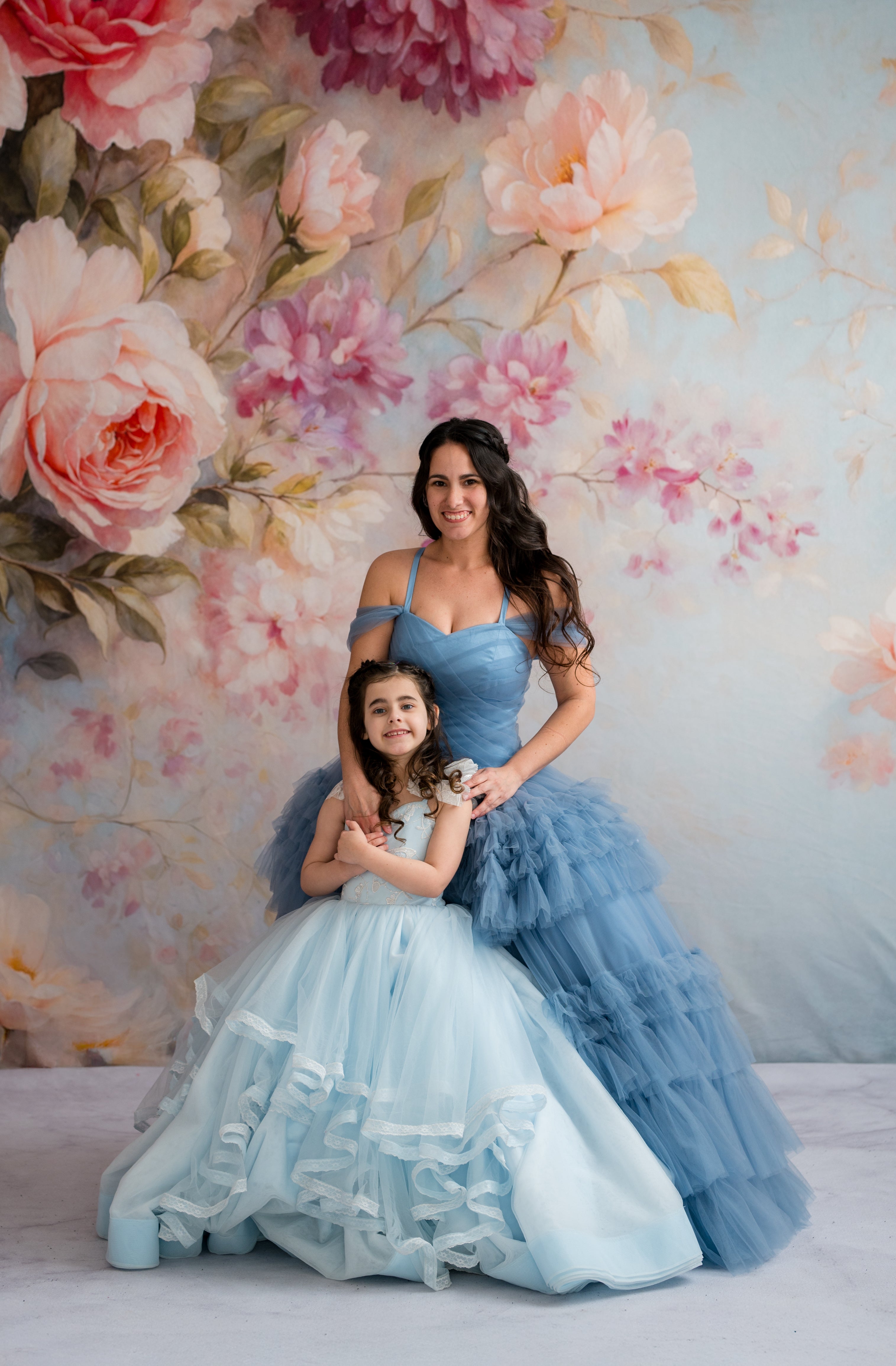 Two women in elegant dresses standing against a floral wall backdrop