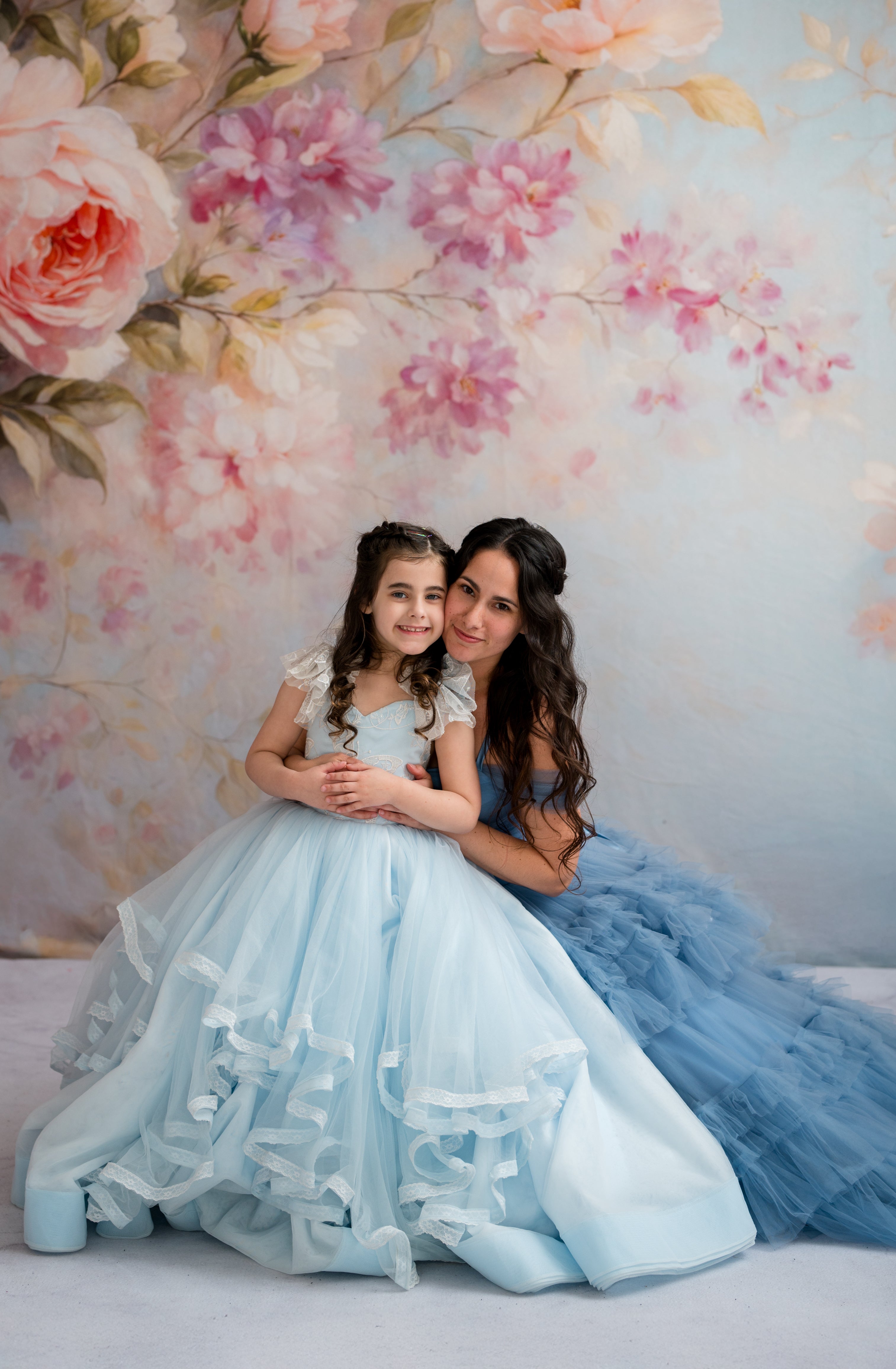 Two women in light blue dresses sitting together against a floral wall.