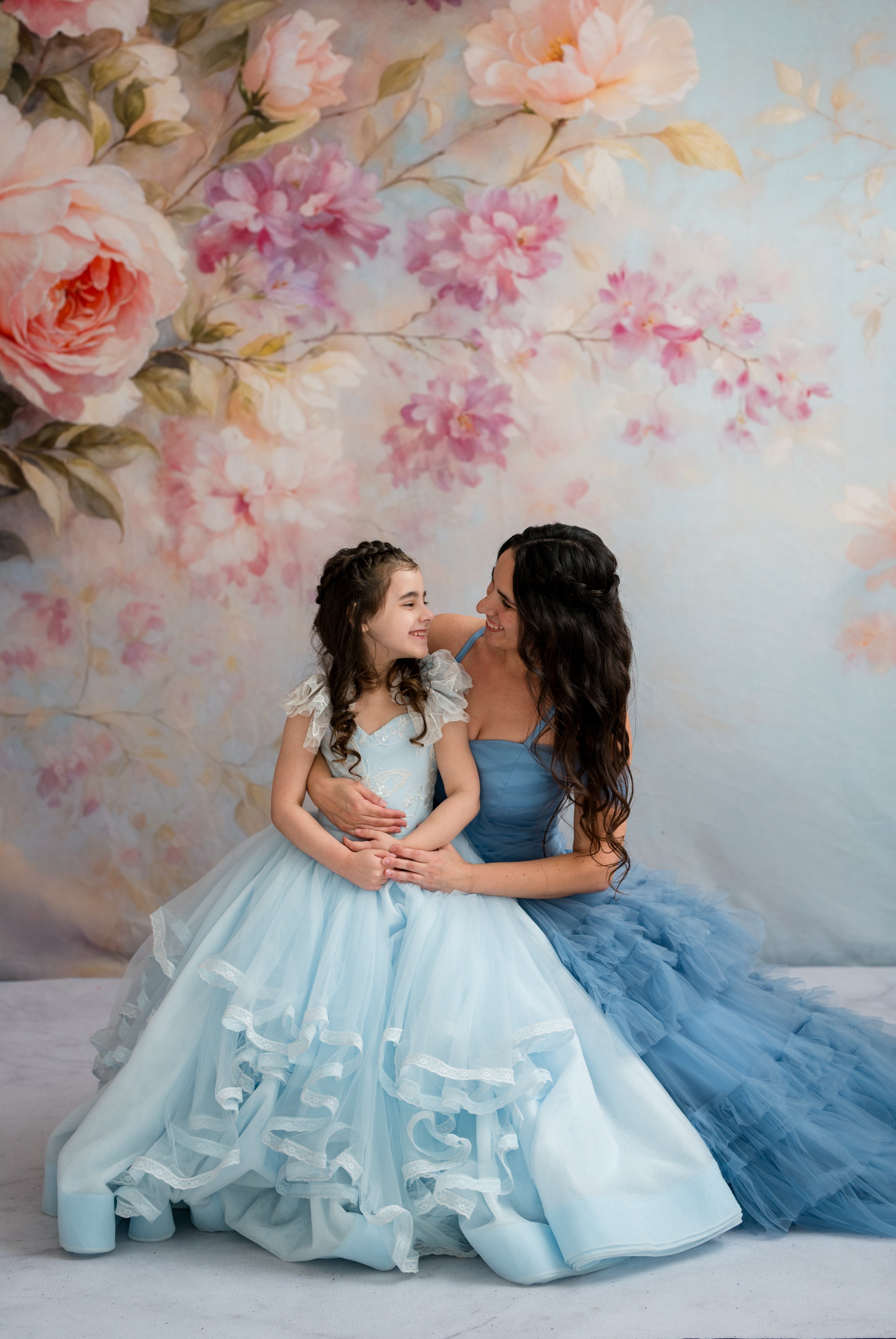 Two women in light blue and blue dresses sitting against a floral wall.