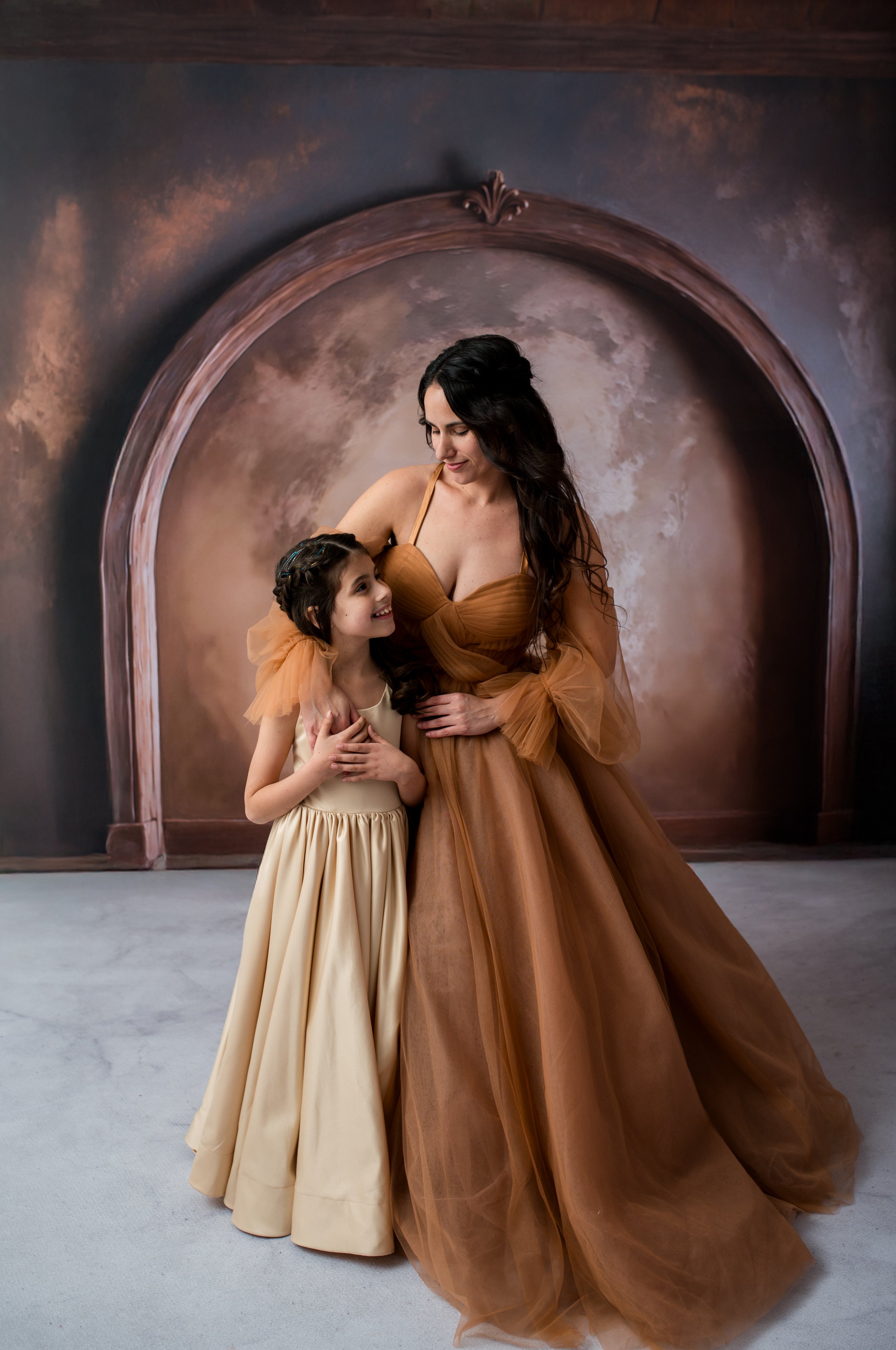 Two women in elegant dresses standing in front of an archway with a textured wall.