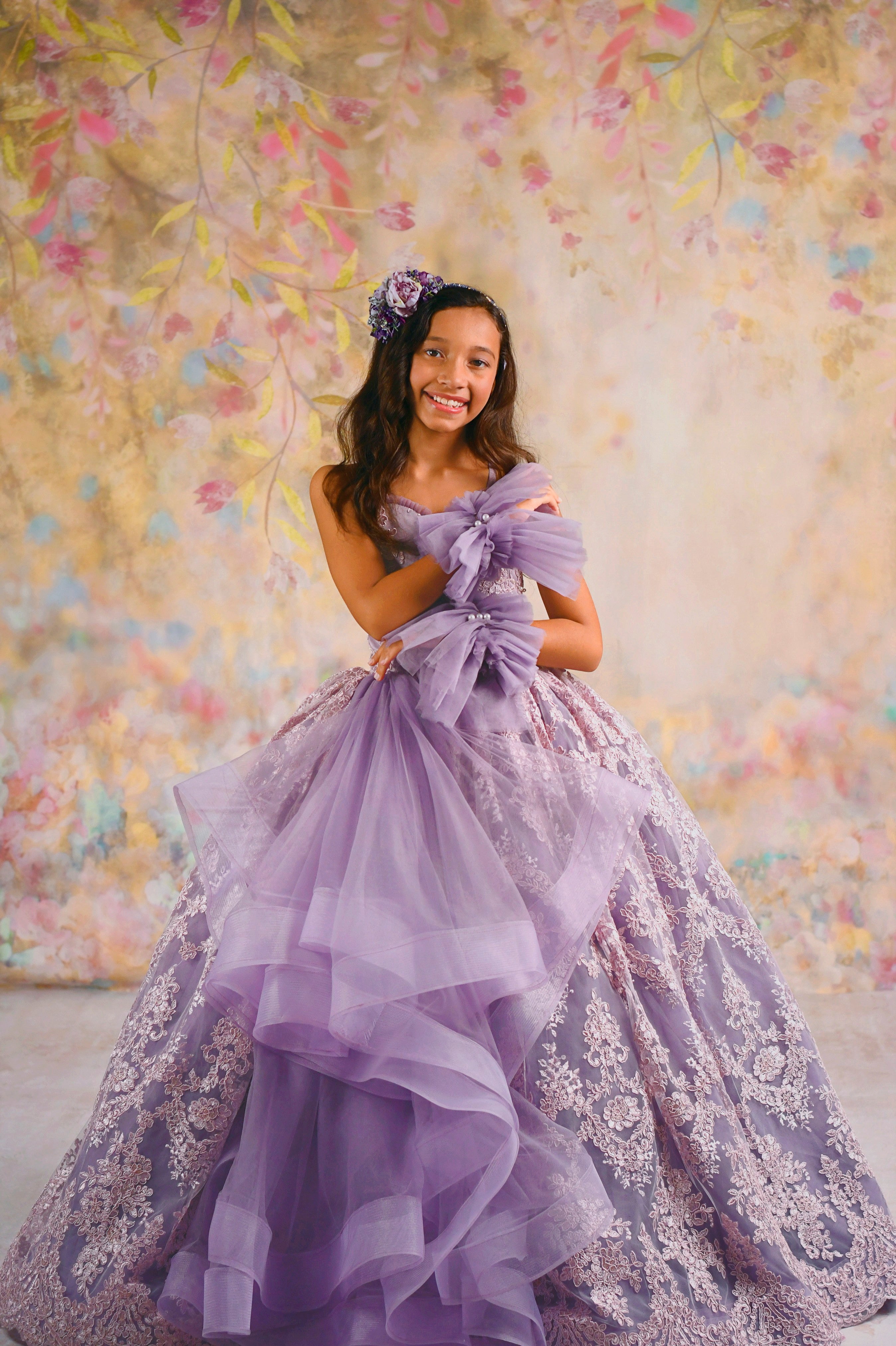 A young girl in a floor-length lavender gown stands in front of a soft floral backdrop. The dress features intricate lace embroidery, layers of voluminous tulle, and oversized bow accents. She is smiling gracefully, with a matching floral headpiece, highlighting the gown's elegant and whimsical design.